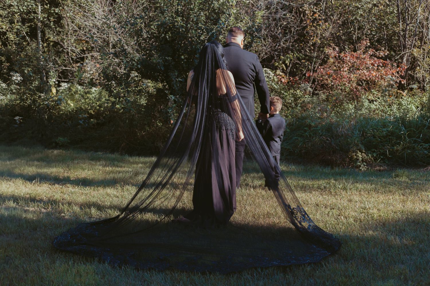 Bride and groom with dramatic long black veil walking through a green outdoor setting.