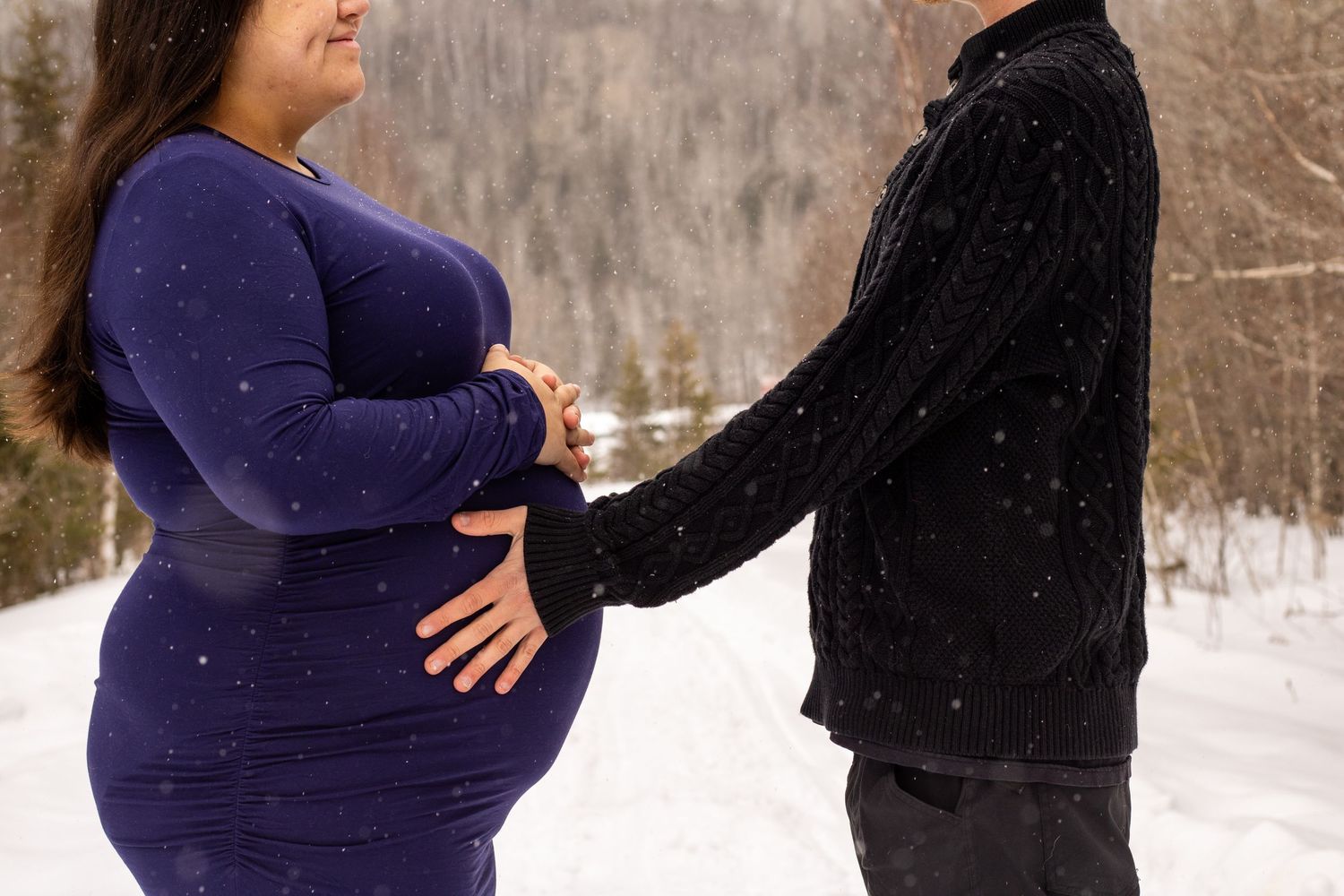 Couple sharing tender moment outdoors in snow, hands on pregnant belly in winter.