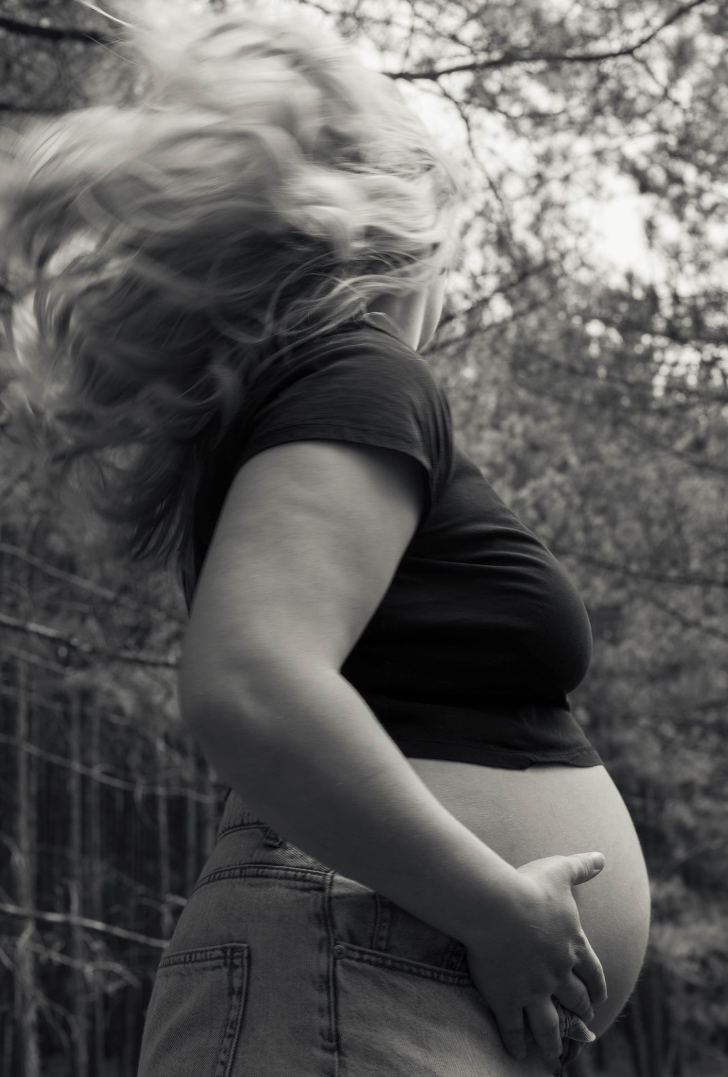 Black and white photo of pregnant woman outdoors, hair flowing, showing baby bump in crop top and jeans.