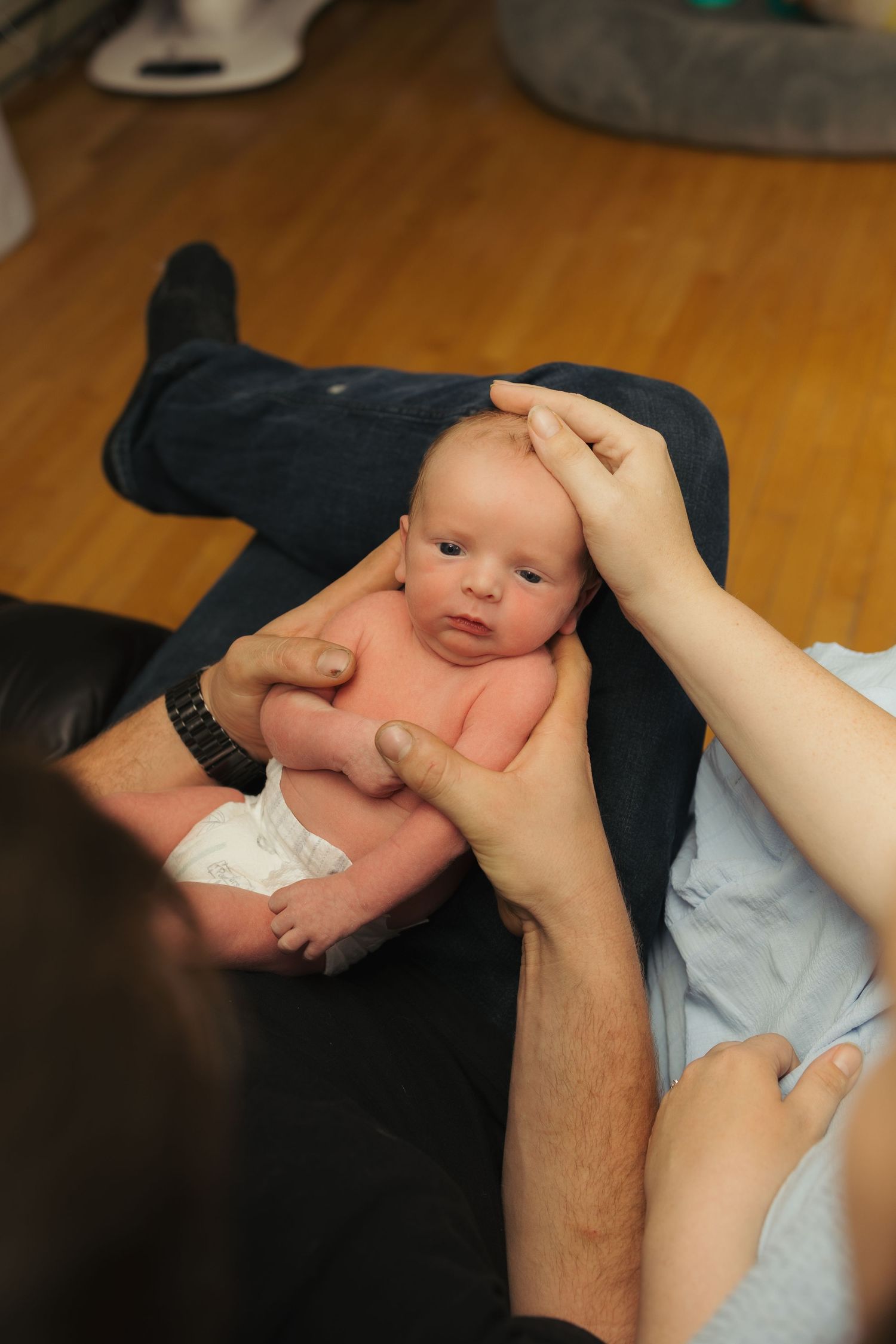 Newborn baby being held by two adults on a wooden floor, wearing only a diaper, looking alert and calm.