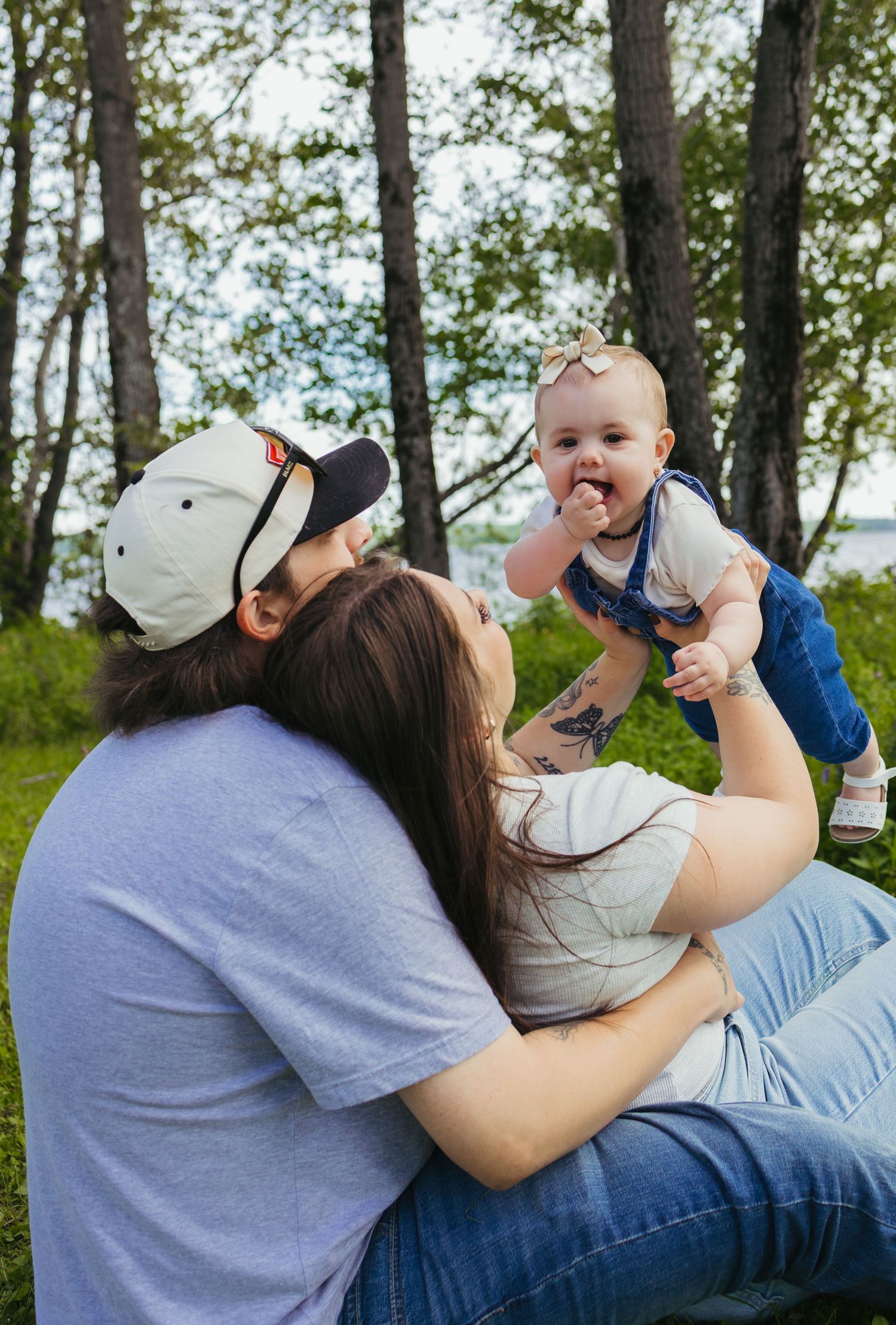 Young couple sitting outdoors in a park, joyfully holding a smiling baby in denim overalls among green trees.