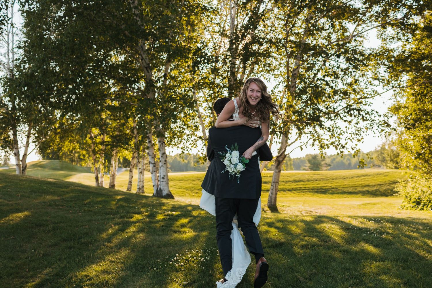 Groom carrying laughing bride outdoors among birch trees on sunny wedding day in lush green park setting.