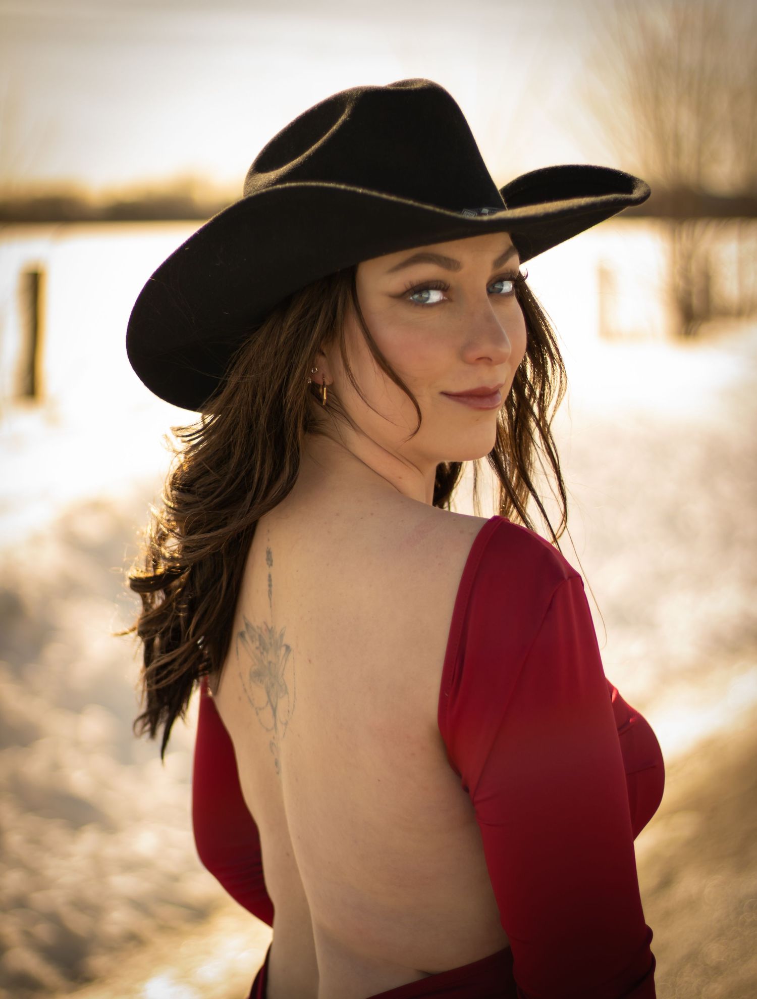 A woman in a red backless dress and black cowboy hat poses outdoors in a snowy winter landscape.