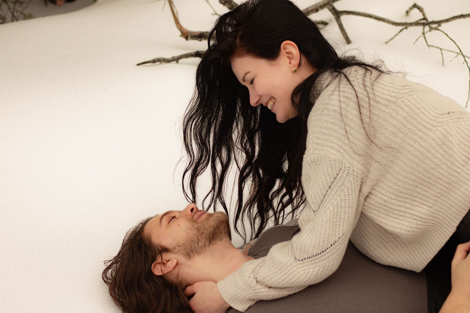 A joyful couple laughs together lying in the snow, surrounded by bare winter branches.