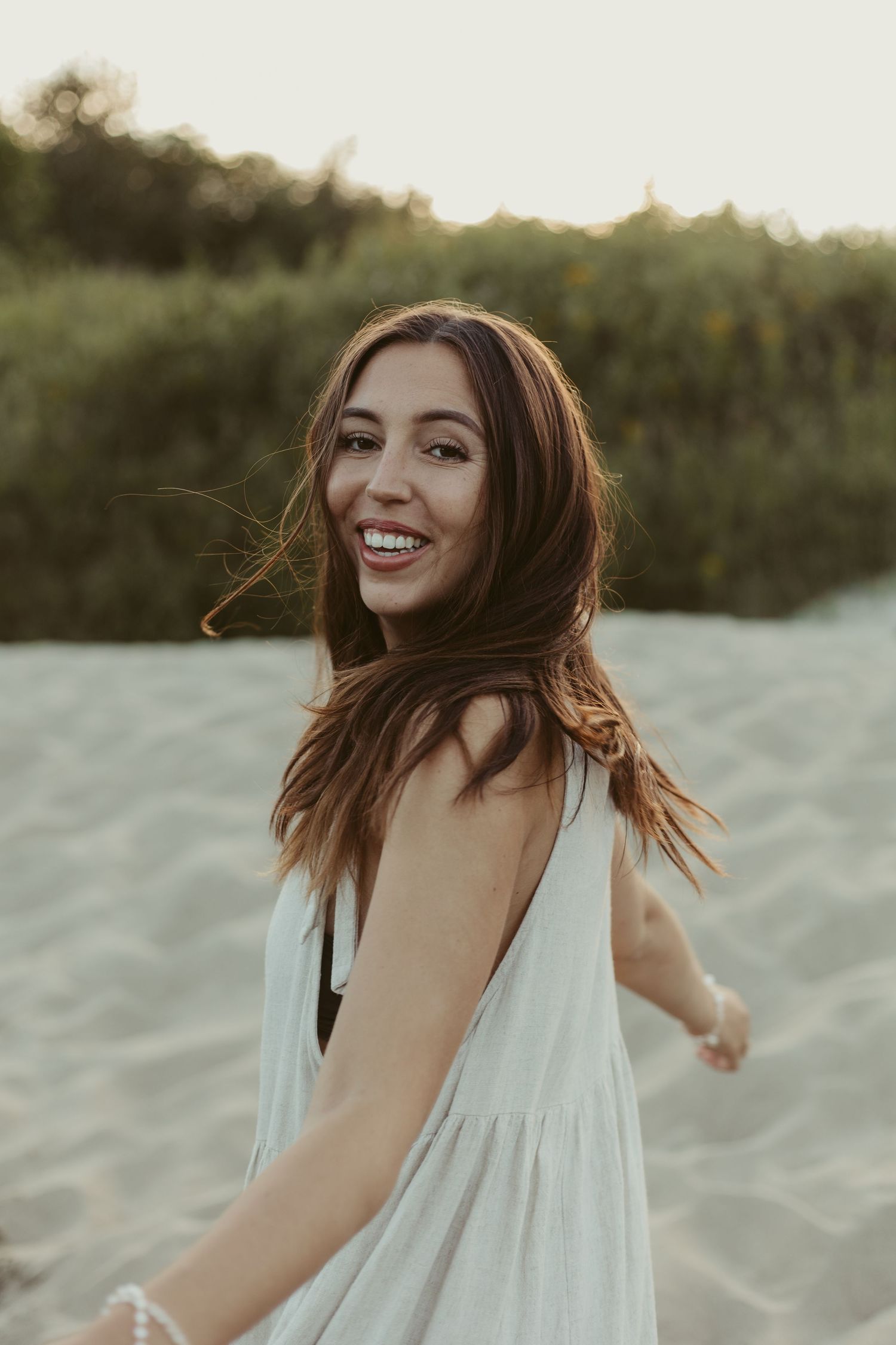 A smiling brunette woman in a white backless dress twirls on a sandy beach at sunset.