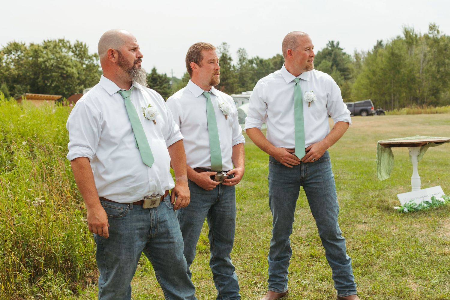 Three groomsmen in white shirts and green ties stand together at an outdoor wedding venue.
