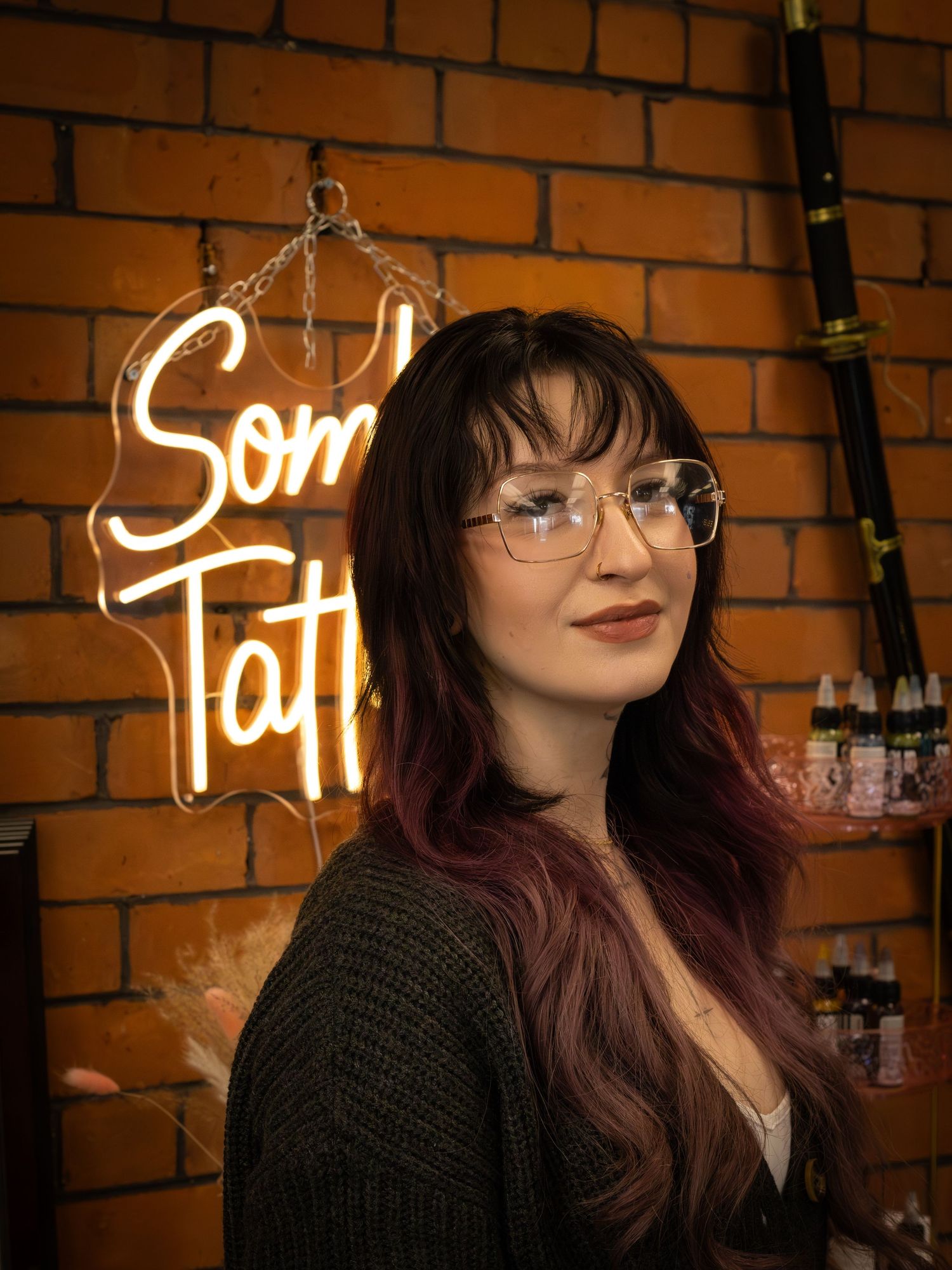 Tattooed woman with glasses posing before a neon tattoo shop sign on brick wall.