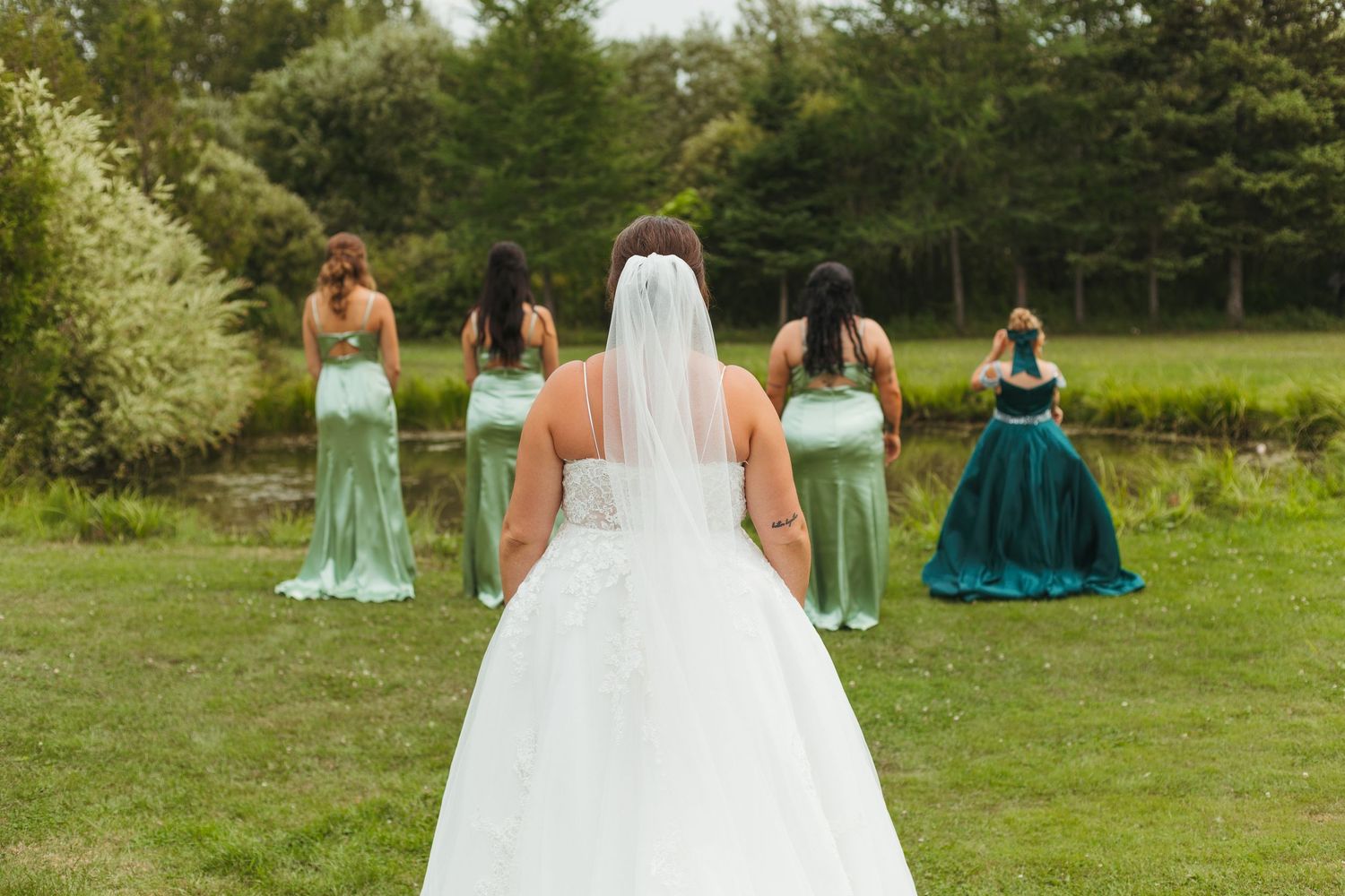 A bride in a white gown and veil stands before her bridesmaids in green dresses outdoors.
