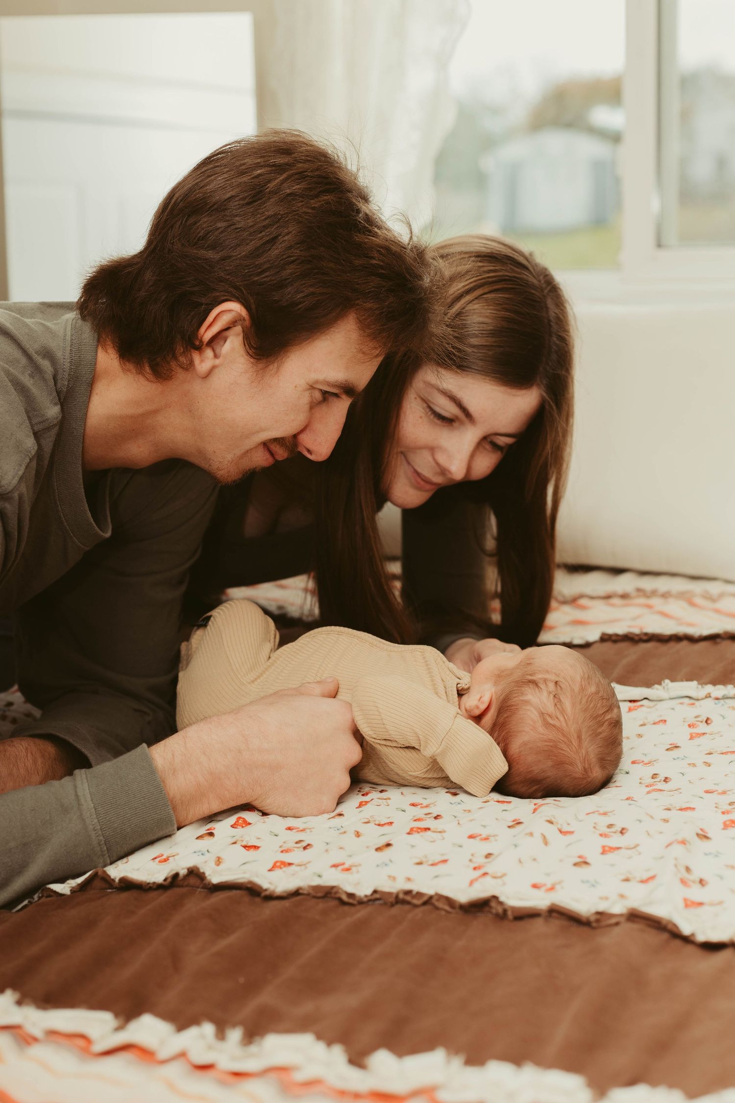 Smiling new parents lying on a bed admiring their peaceful newborn baby on a quilt.