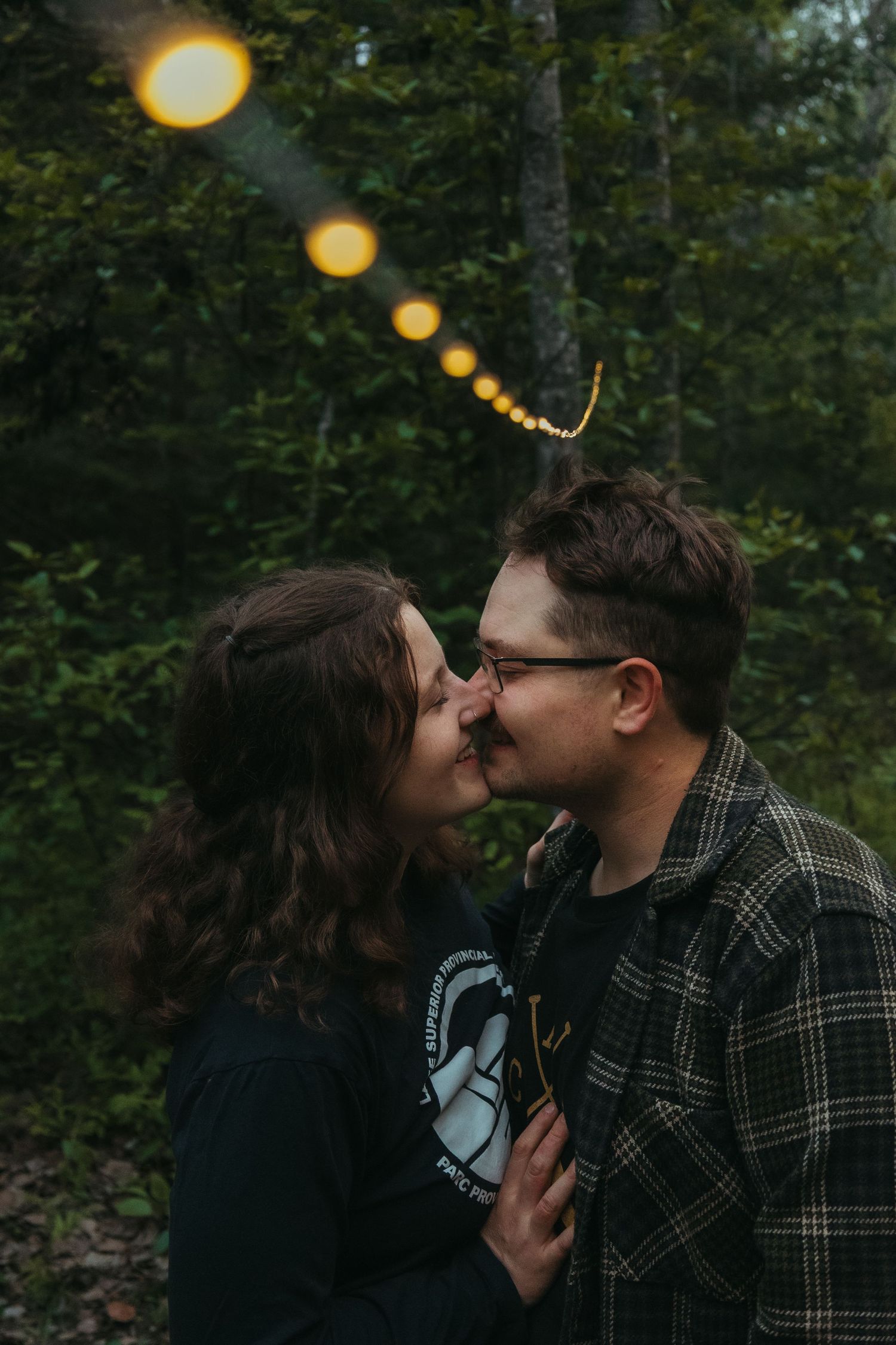 Couple sharing a romantic kiss under string lights in a lush forest at dusk.