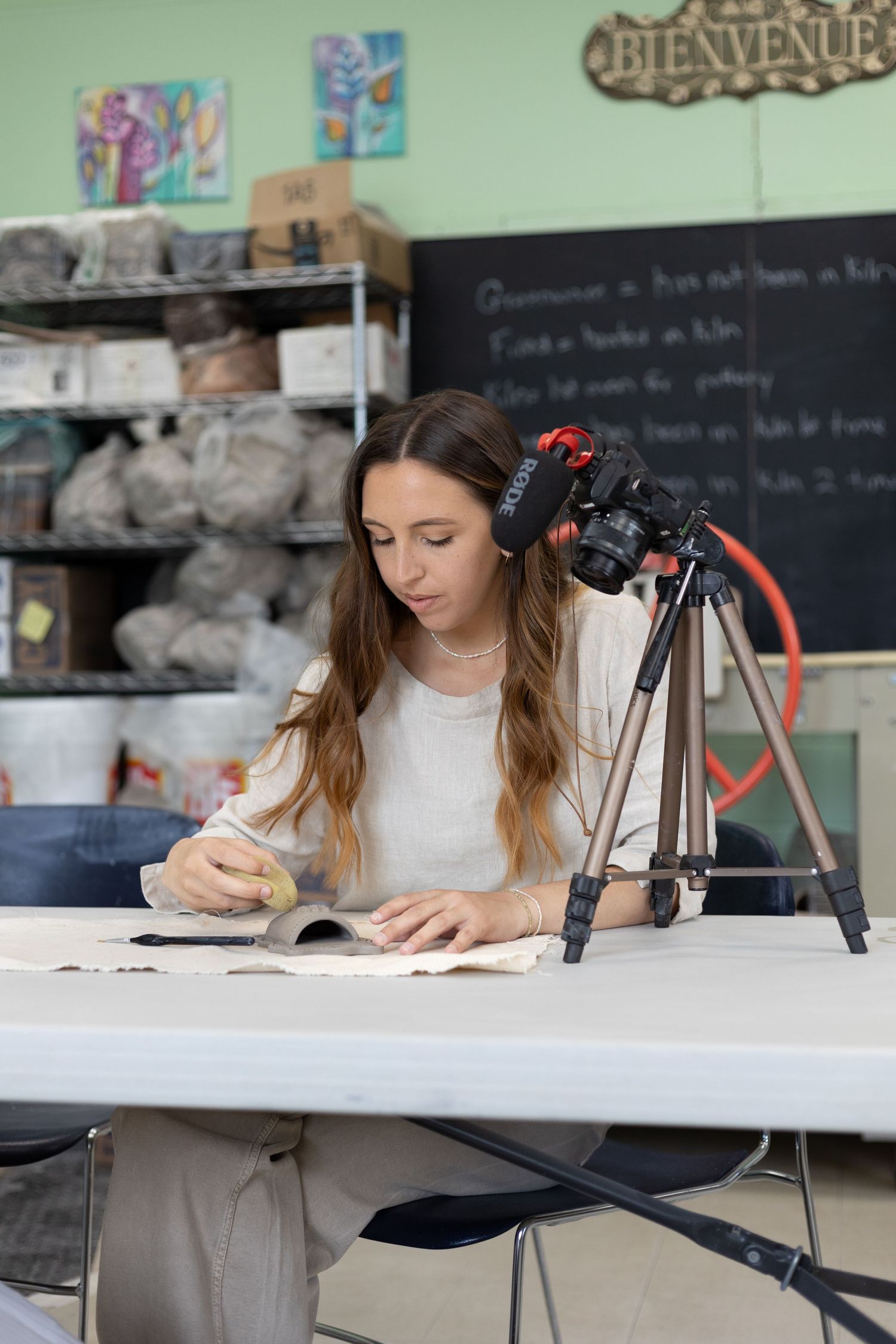 Young woman working on pottery at a white table with a camera on tripod in an art studio classroom.