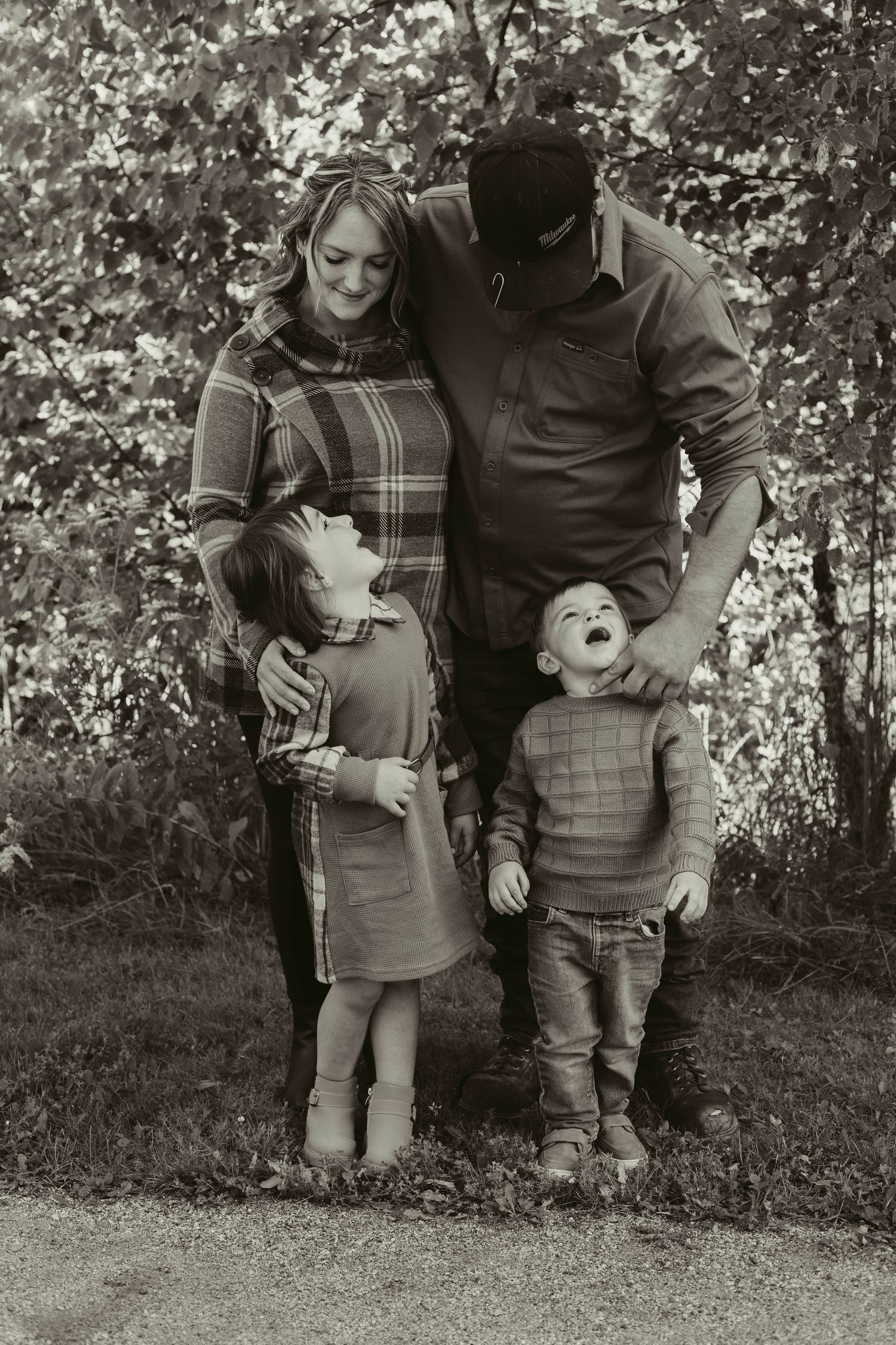 A happy family of four poses together outdoors in a black and white portrait among autumn foliage.