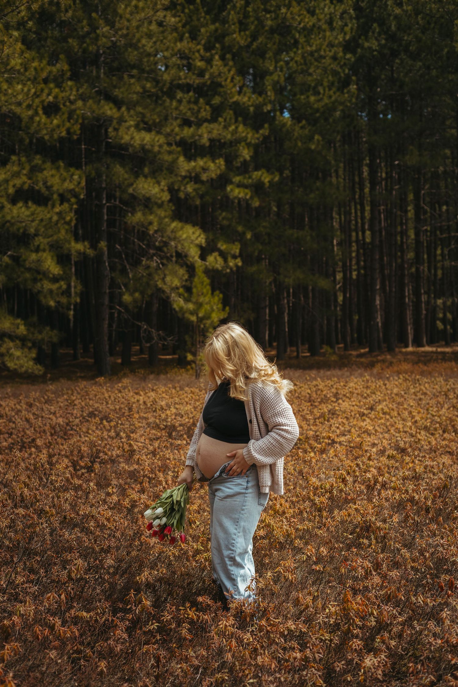 Pregnant woman in jeans and pink cardigan holding tulips in an autumn forest clearing with tall pine trees.