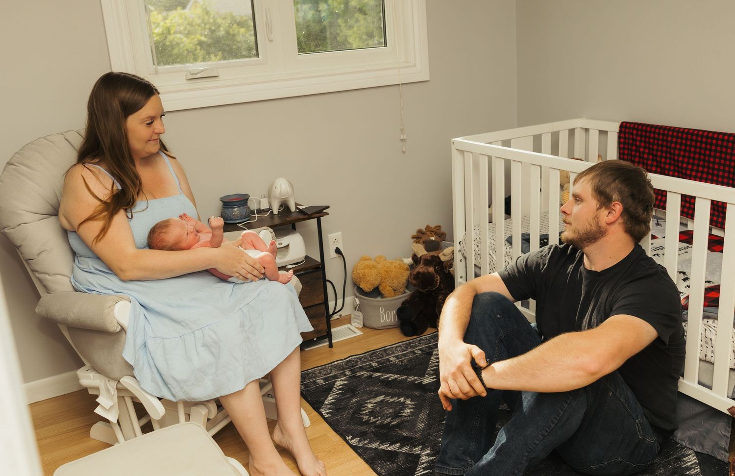 New parents sitting in nursery room, mother holding newborn baby in rocking chair while father sits on floor nearby.