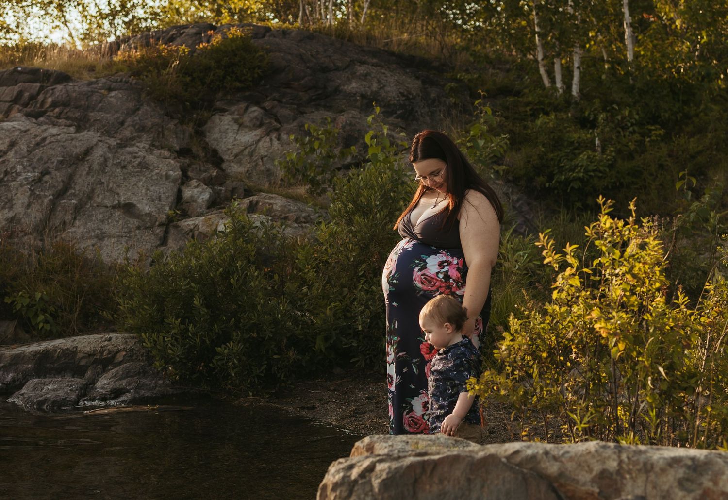 Pregnant woman in floral dress standing on rocks by a river with a young child, surrounded by lush greenery at sunset.