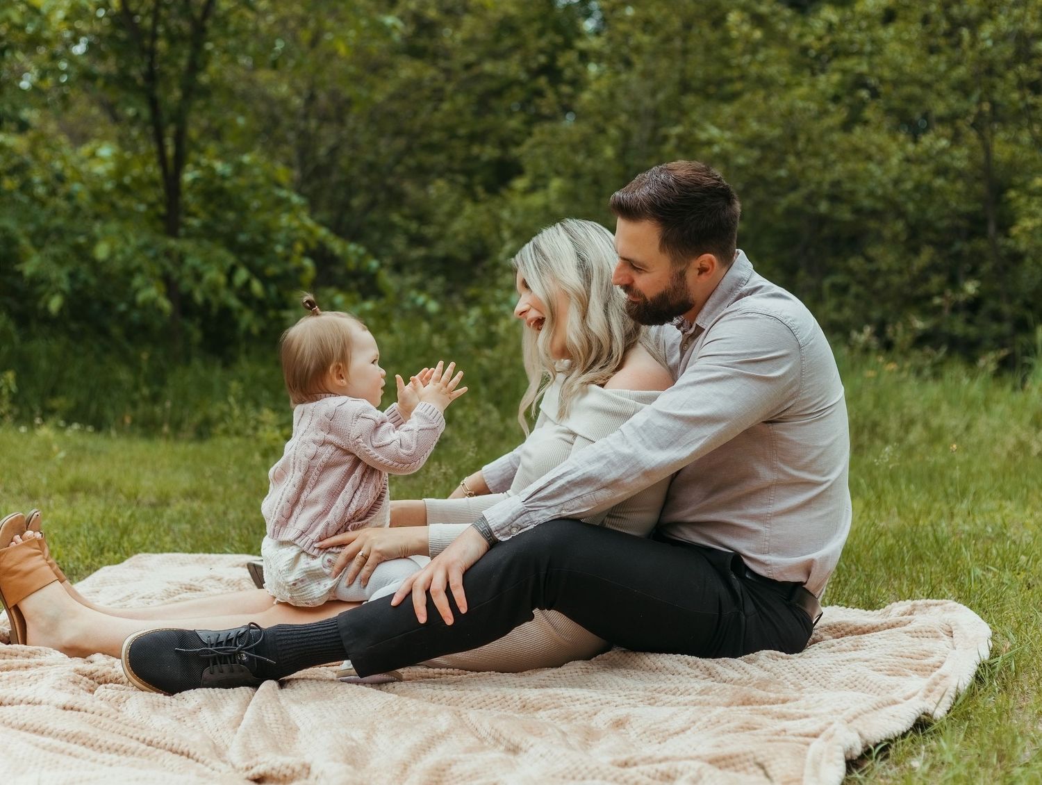 Family sitting on blanket in green park, parents smiling at clapping baby girl on sunny day.