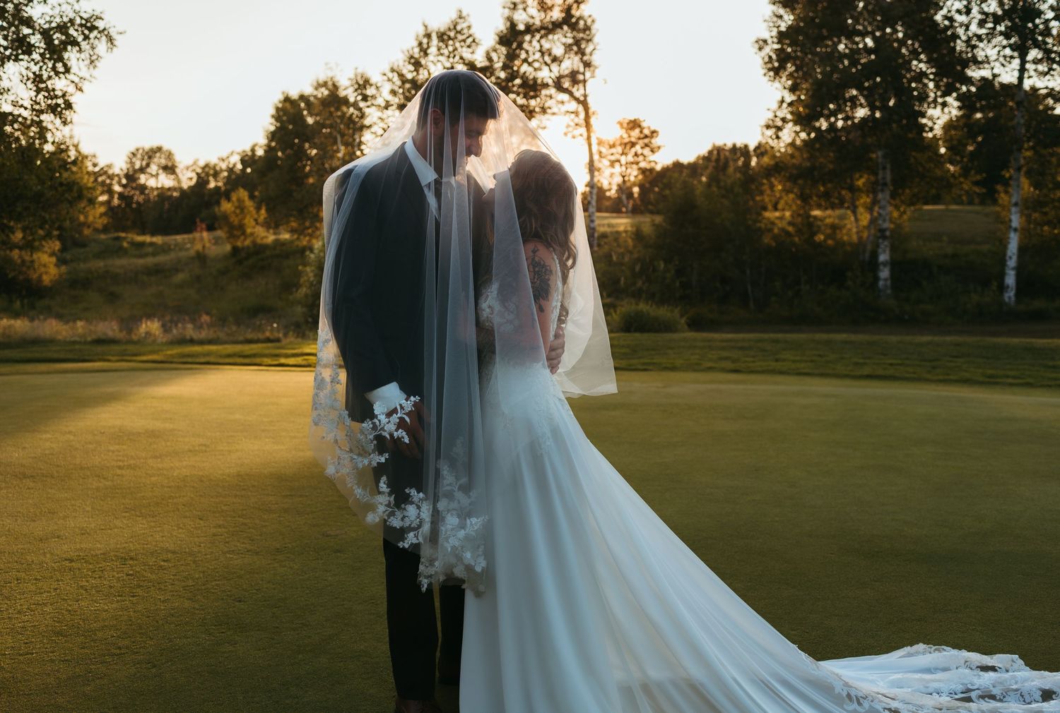 Bride and groom share a romantic kiss under a veil at golden hour on a lush golf course.