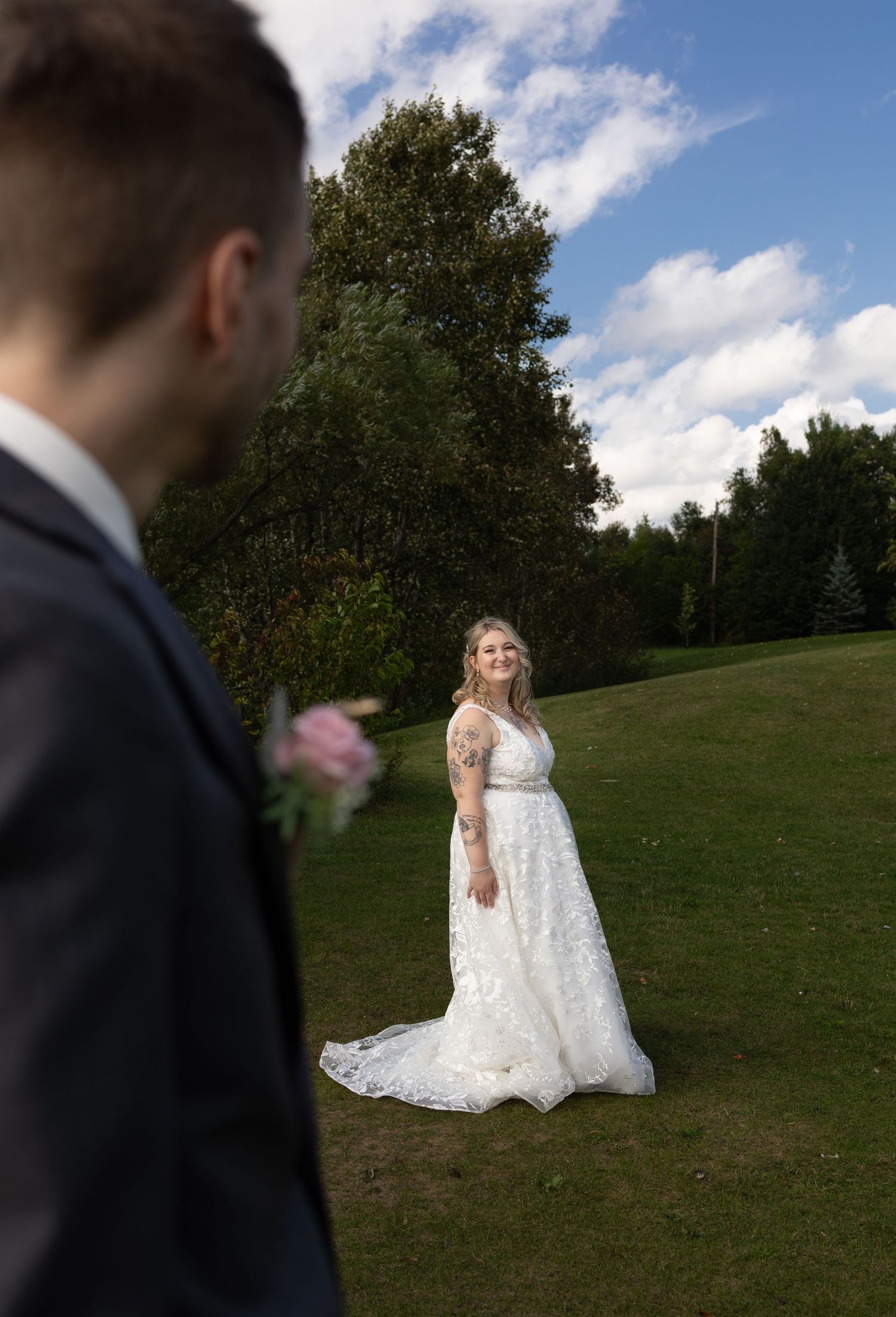 Bride in lace wedding gown smiles at groom outdoors on green lawn with trees and blue sky backdrop.