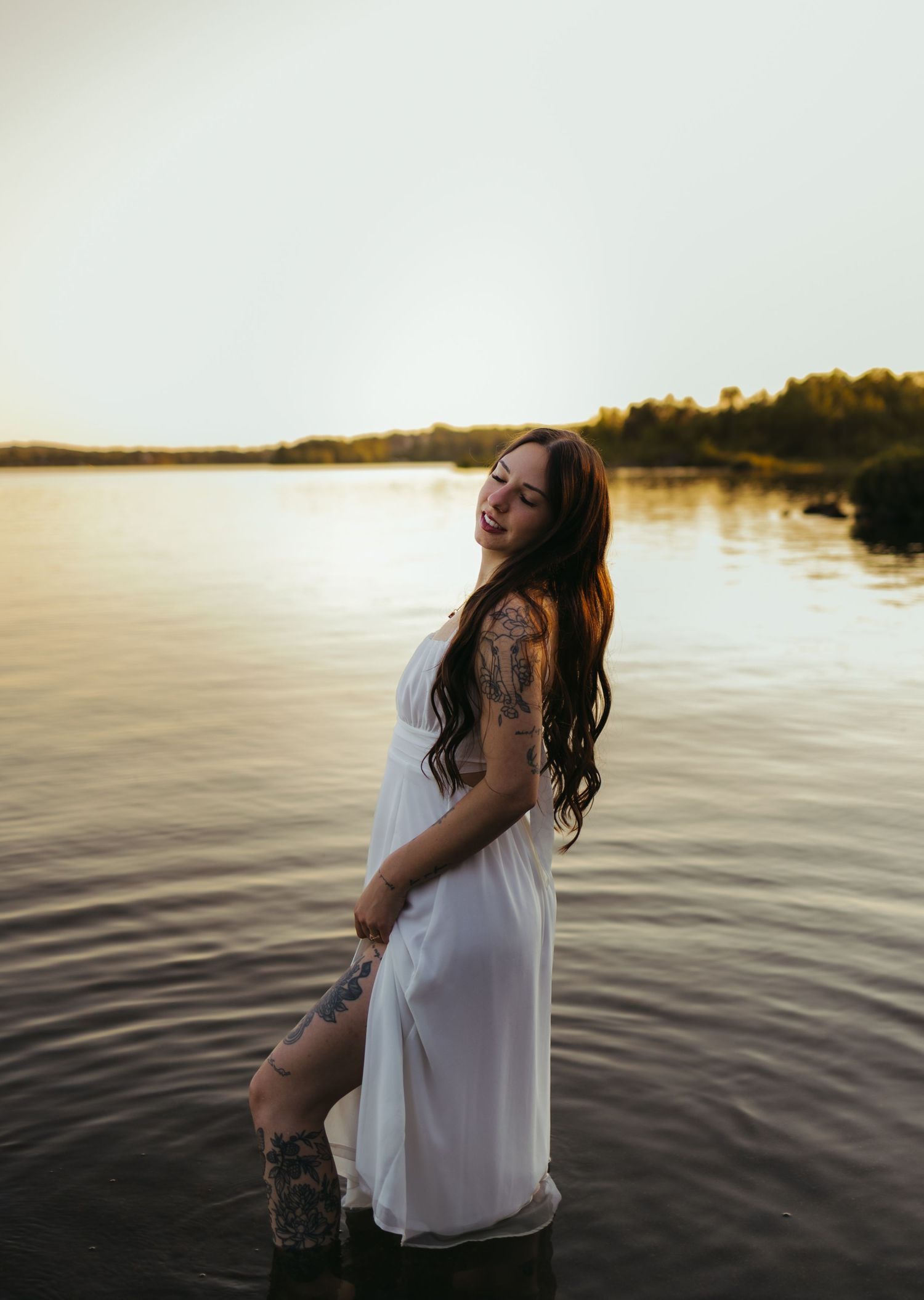 A tattooed woman in a white dress stands in a calm lake at golden hour sunset.