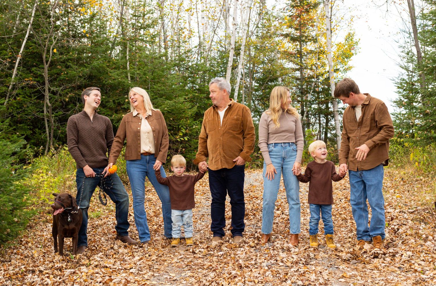 Multi-generational family with dog poses outdoors on fallen autumn leaves surrounded by colorful trees.