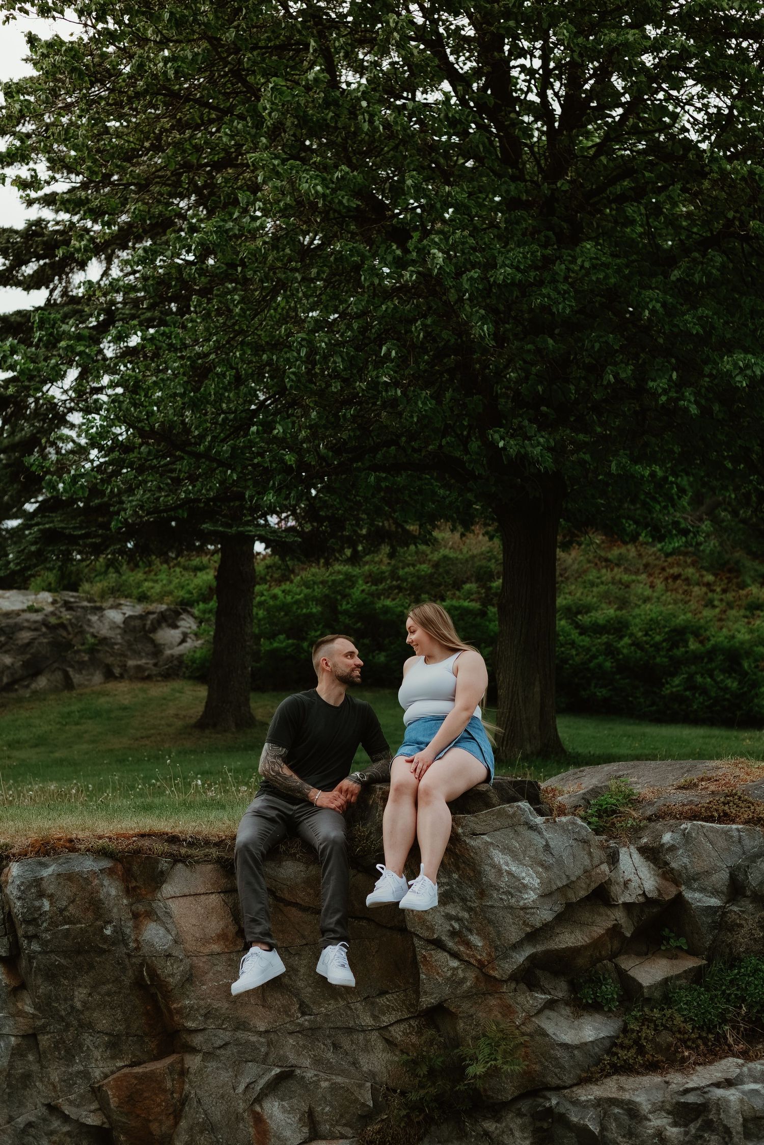 A couple sits on rocky ledge in a lush park, sharing a tender moment surrounded by green trees.