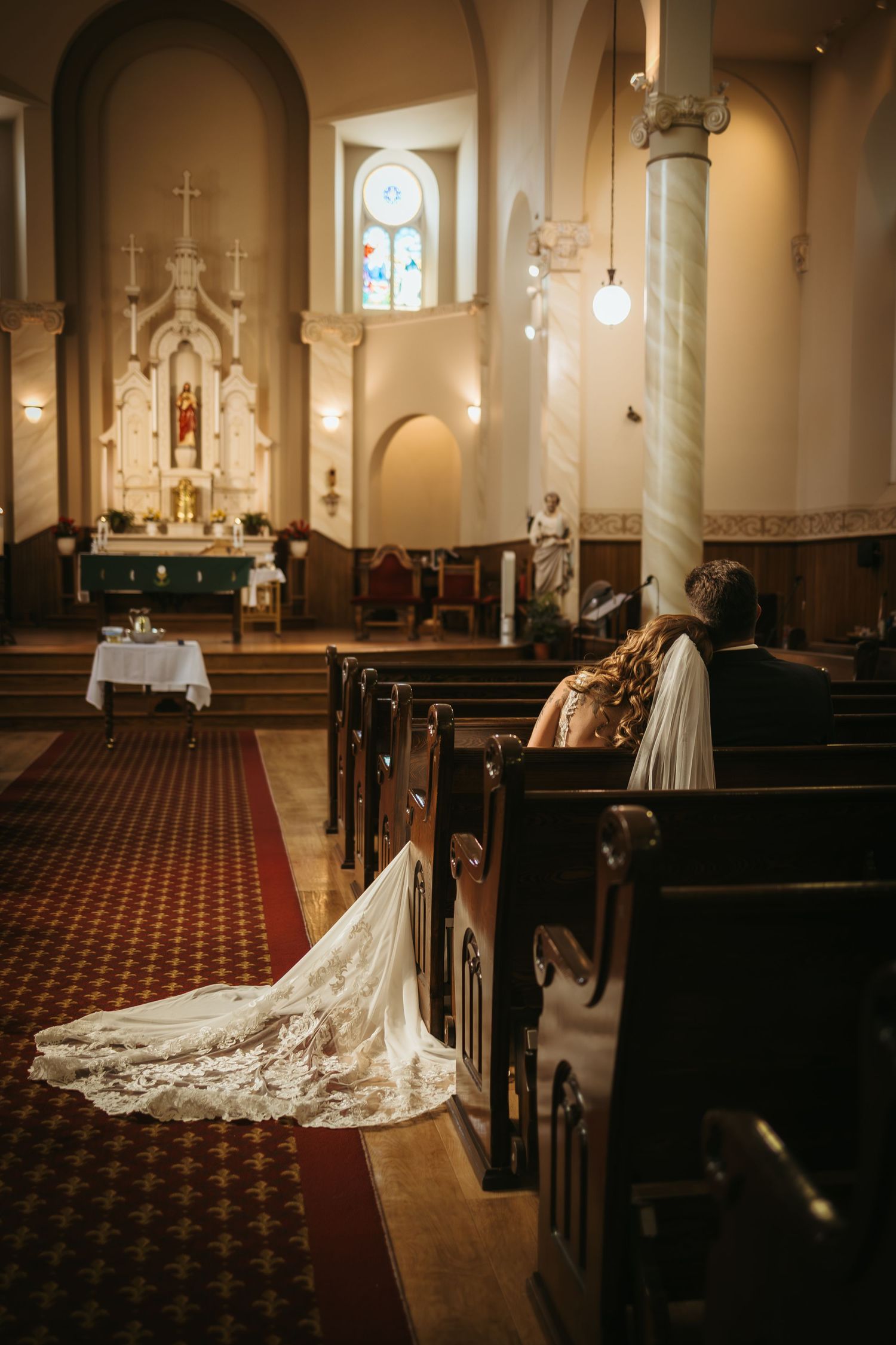 Bride and groom sit in church pews during wedding ceremony, with elegant lace bridal train trailing along the aisle.