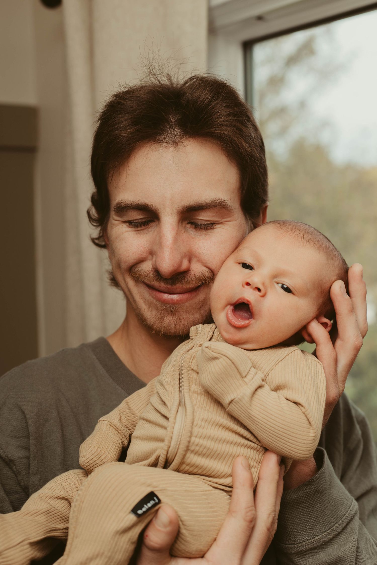 Smiling father holding wide-eyed yawning newborn baby close to his cheek indoors.