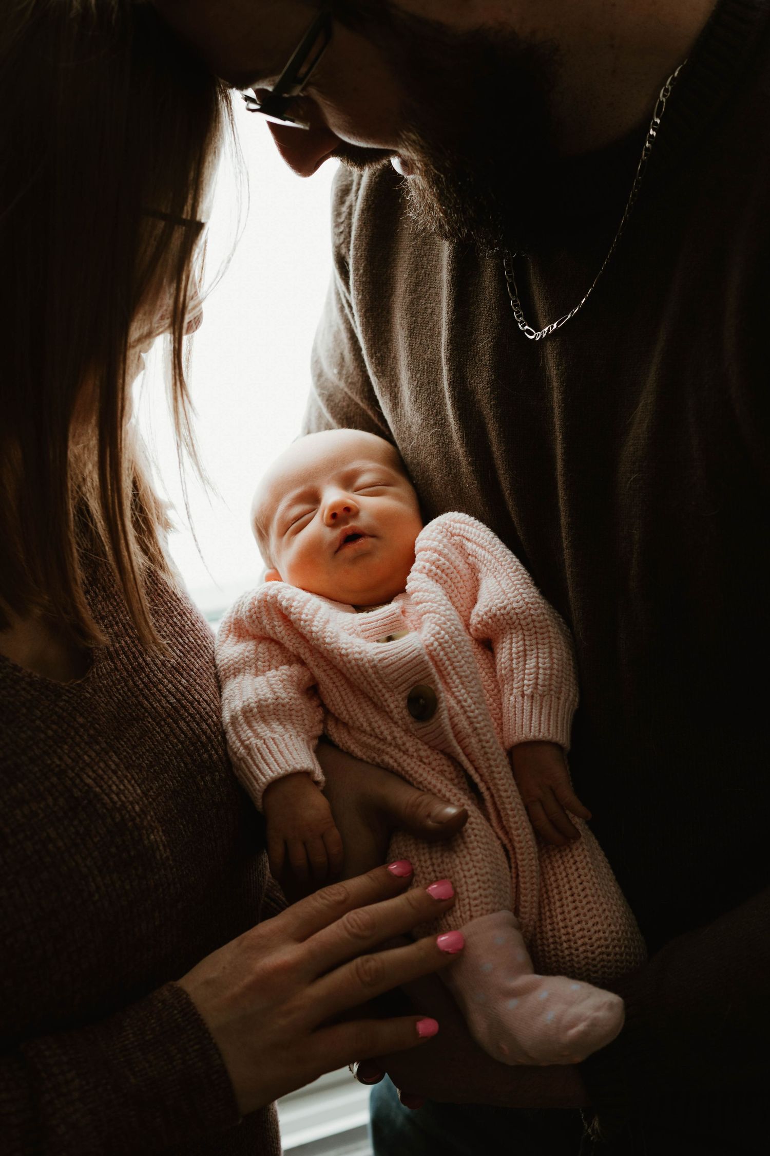 New parents lean in together to admire their sleeping newborn baby dressed in a pink knit outfit.