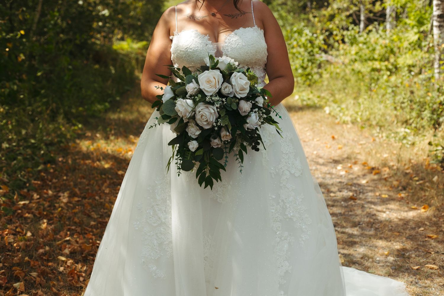 Plus-size bride holding large white rose bouquet in lace wedding gown on forest path.