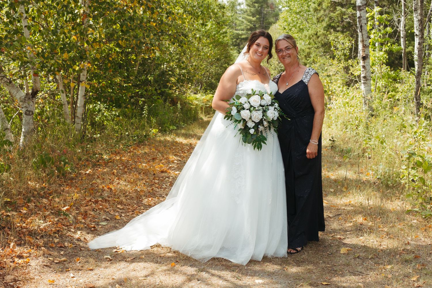 Bride in white wedding gown posing with woman in black dress holding bouquet among birch trees.