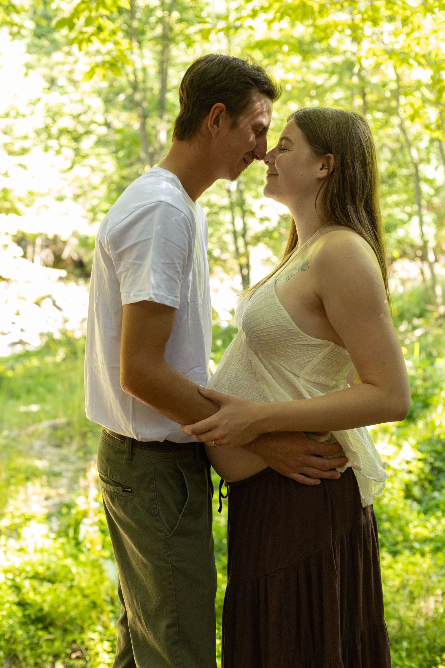 Expecting couple sharing a tender forehead-to-forehead moment in a sunlit green forest.
