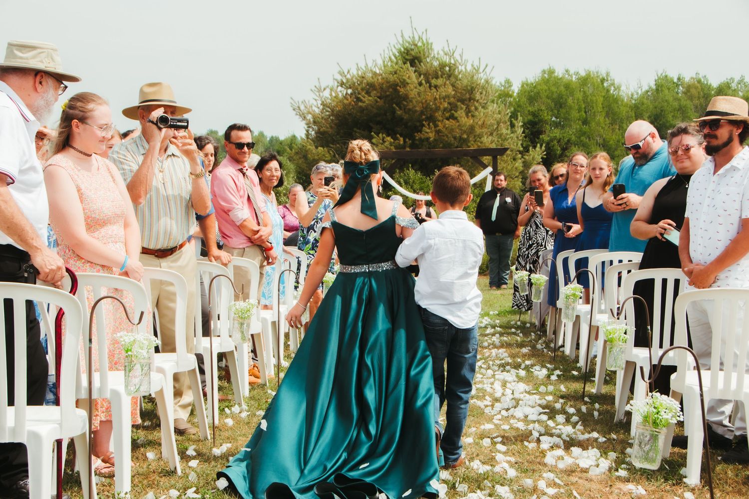 Bride in teal gown walks down outdoor wedding aisle surrounded by cheering guests.
