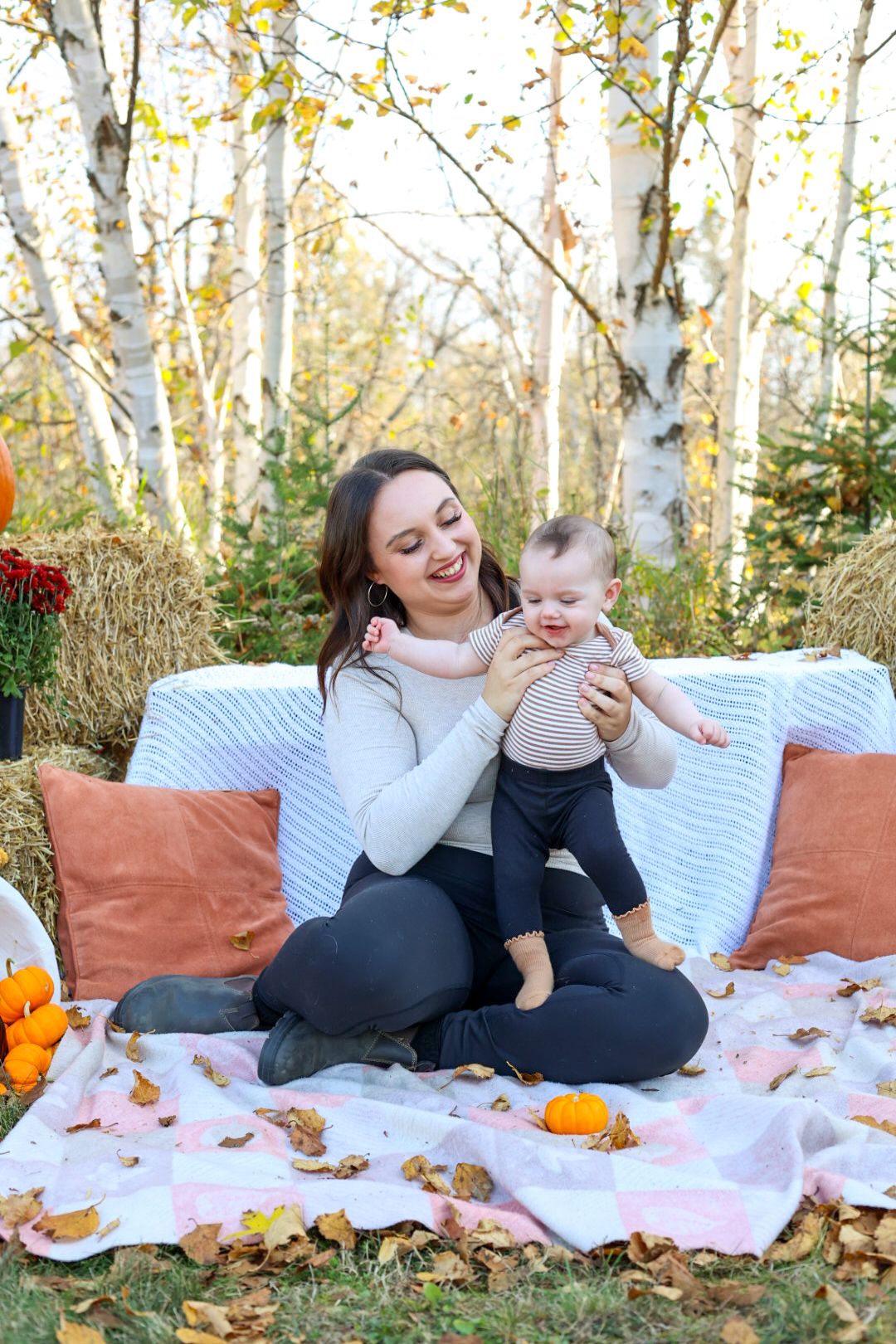 Happy mother holding smiling baby on a picnic blanket surrounded by autumn leaves and birch trees.
