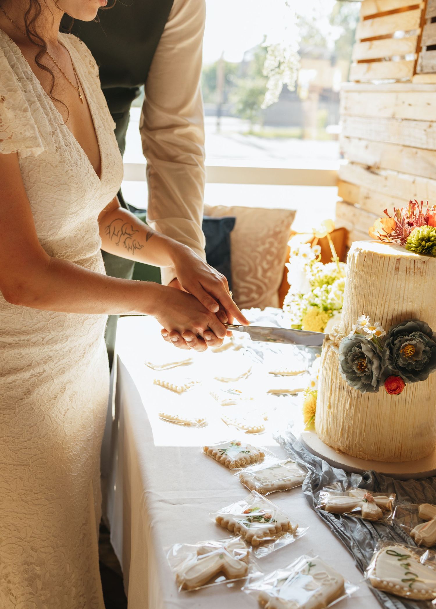 Bride and groom cutting elegant white textured wedding cake with floral topper at outdoor reception table.