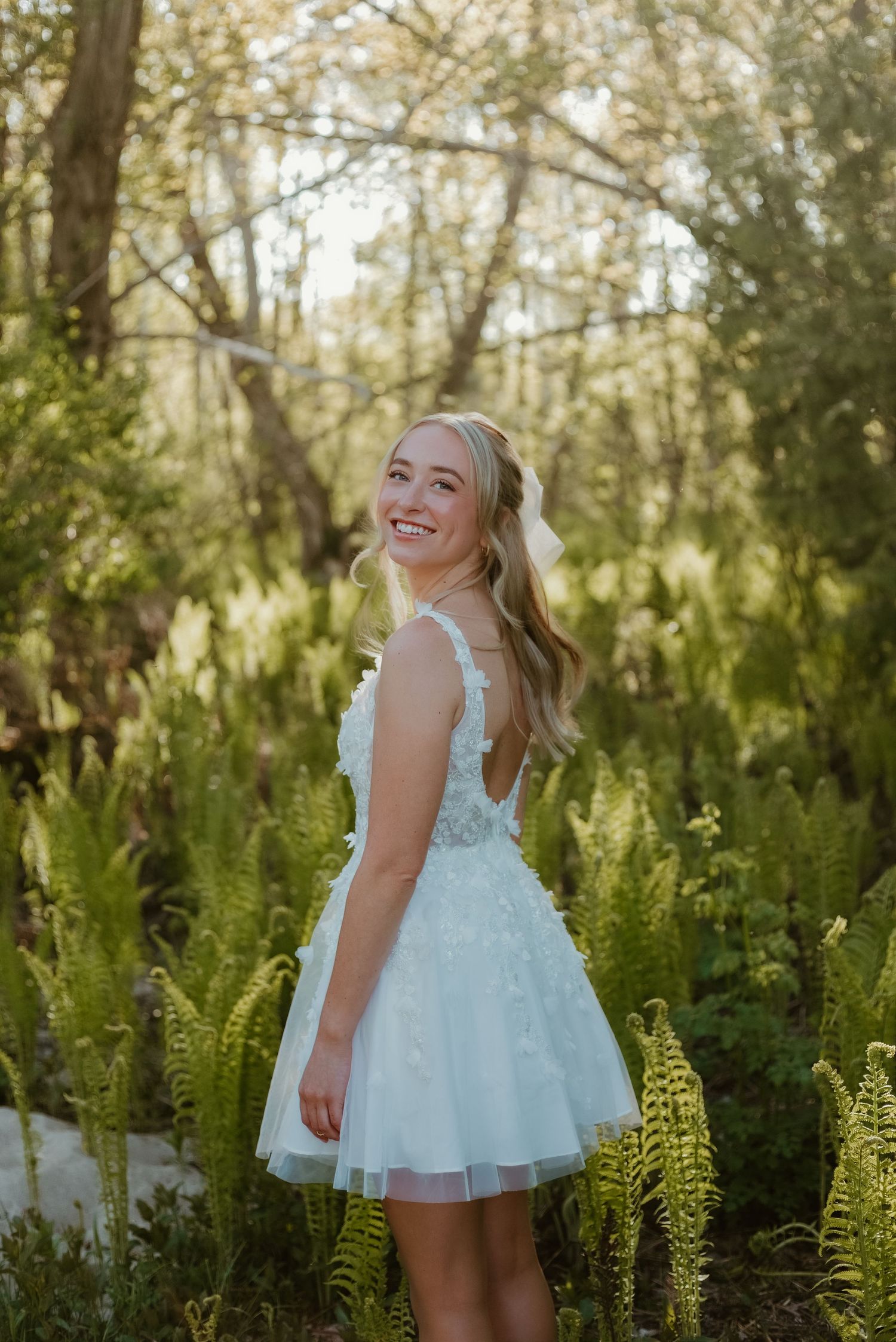 Smiling blonde woman in a light blue floral tulle dress standing among ferns in a sunlit forest.