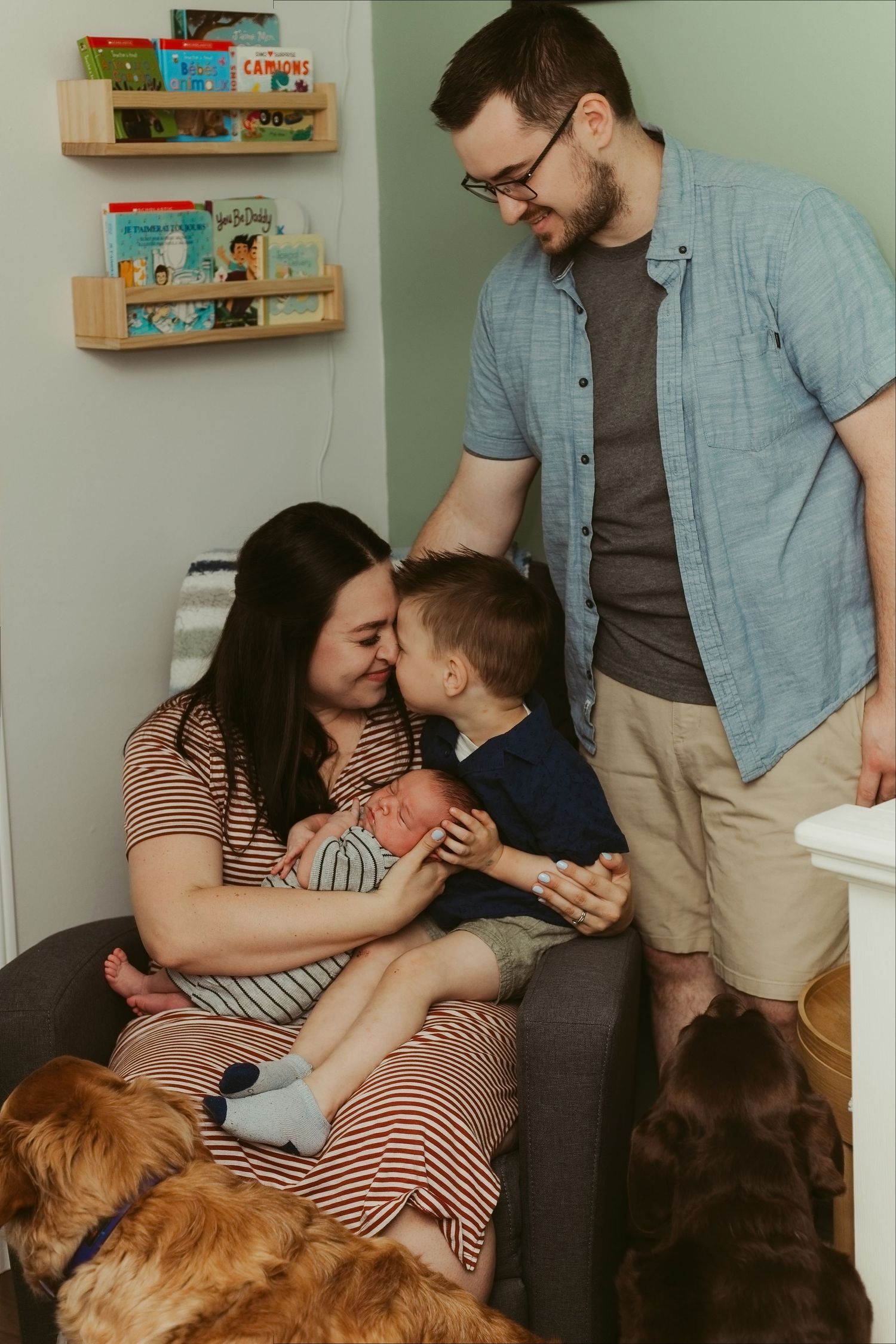 Family with newborn baby, toddler, and golden retriever gathered in a nursery with book shelves.