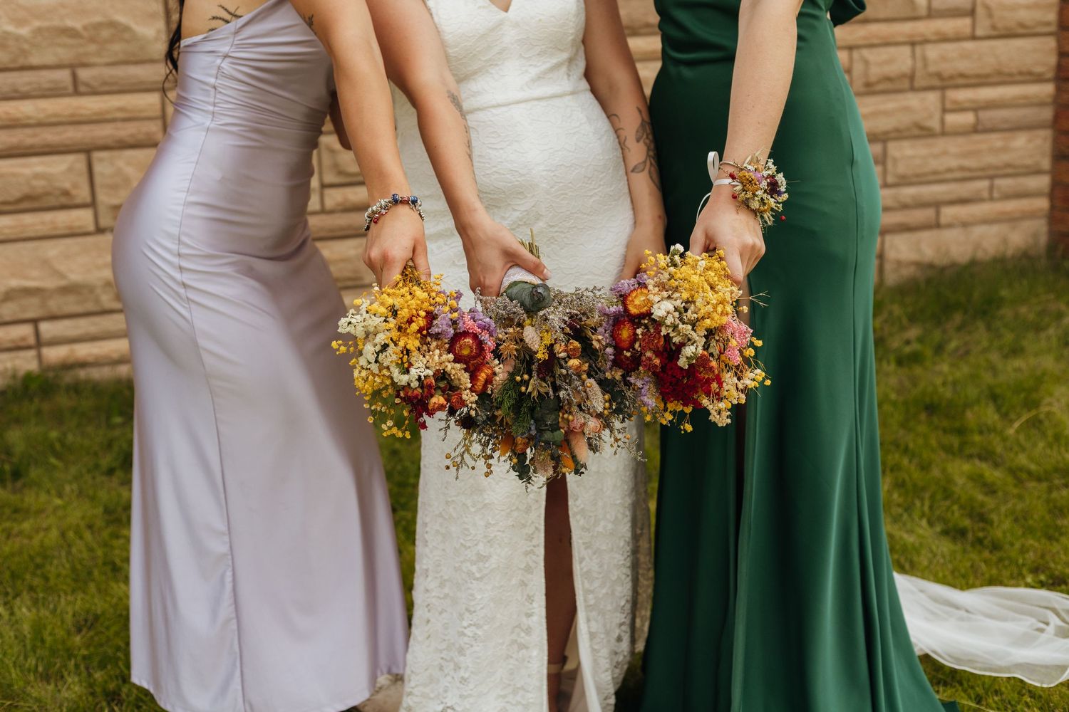 Bride and two bridesmaids in lavender and green gowns holding colorful wildflower bouquets outdoors.