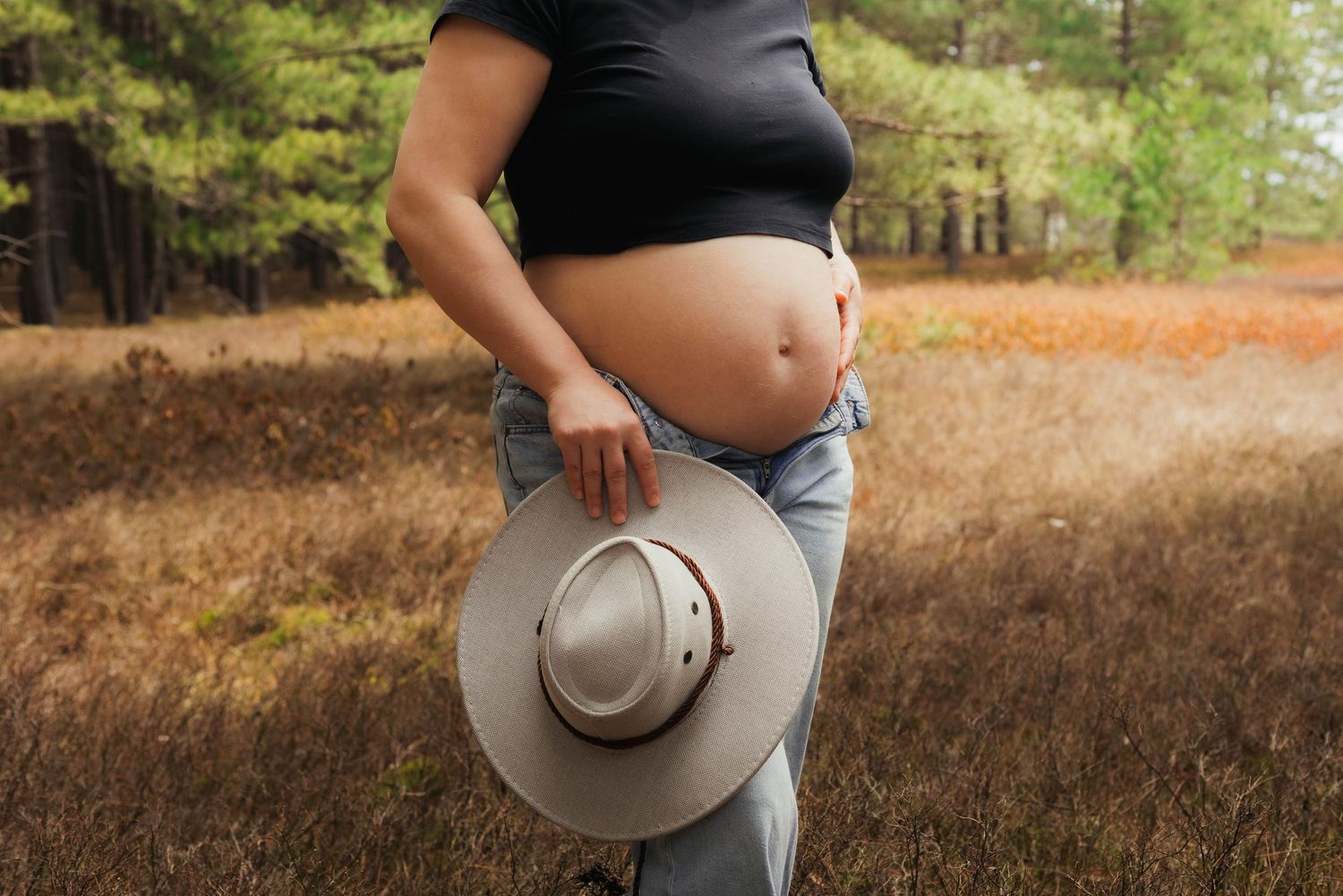 Close-up of pregnant woman holding cowboy hat with tulips tucked in jeans waistband outdoors.