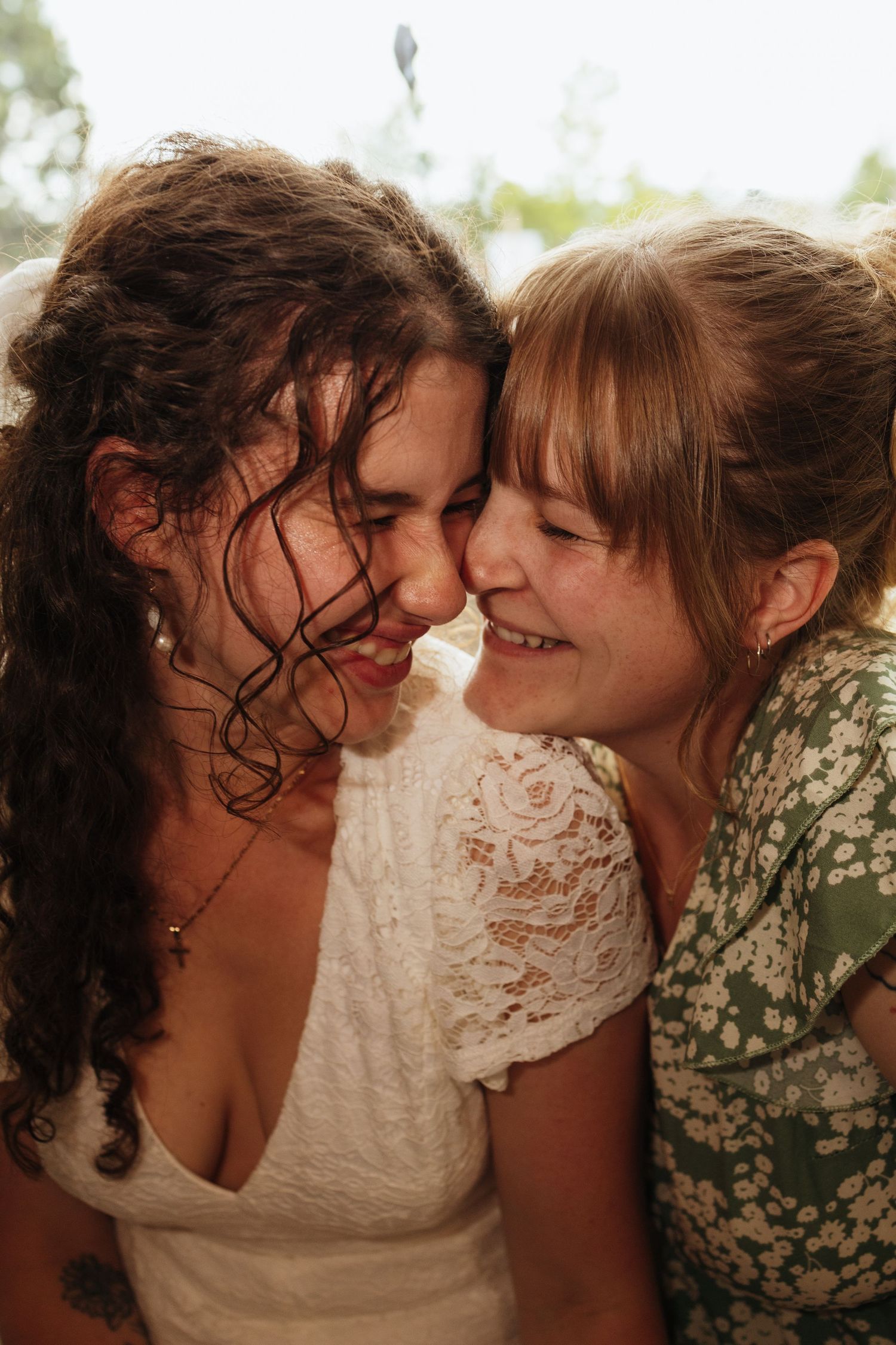 Two women smiling and laughing together, foreheads touching, one in white lace dress, other in green floral top.