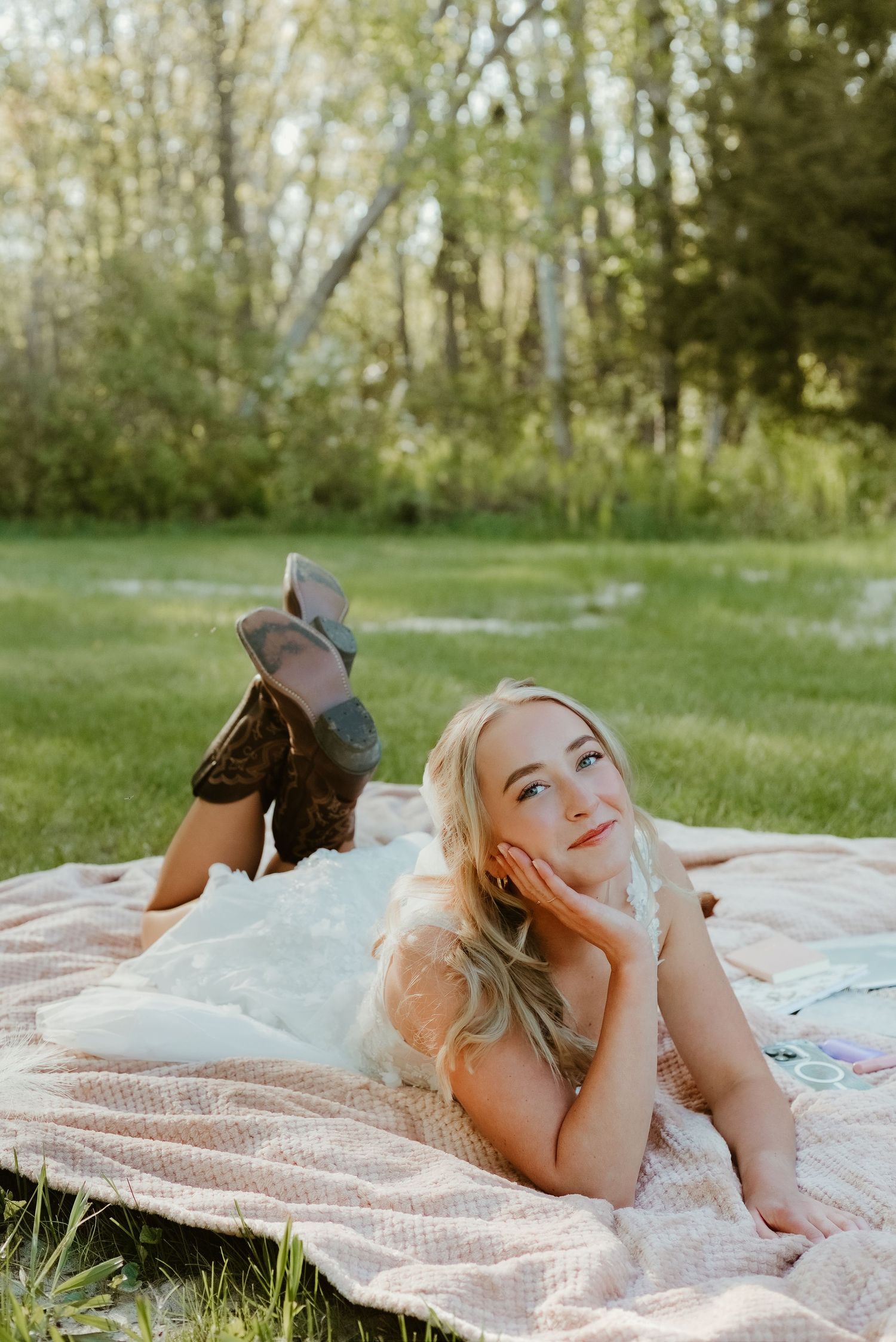 Blonde woman in white dress and cowboy boots lying on a blanket outdoors in a lush green park setting.