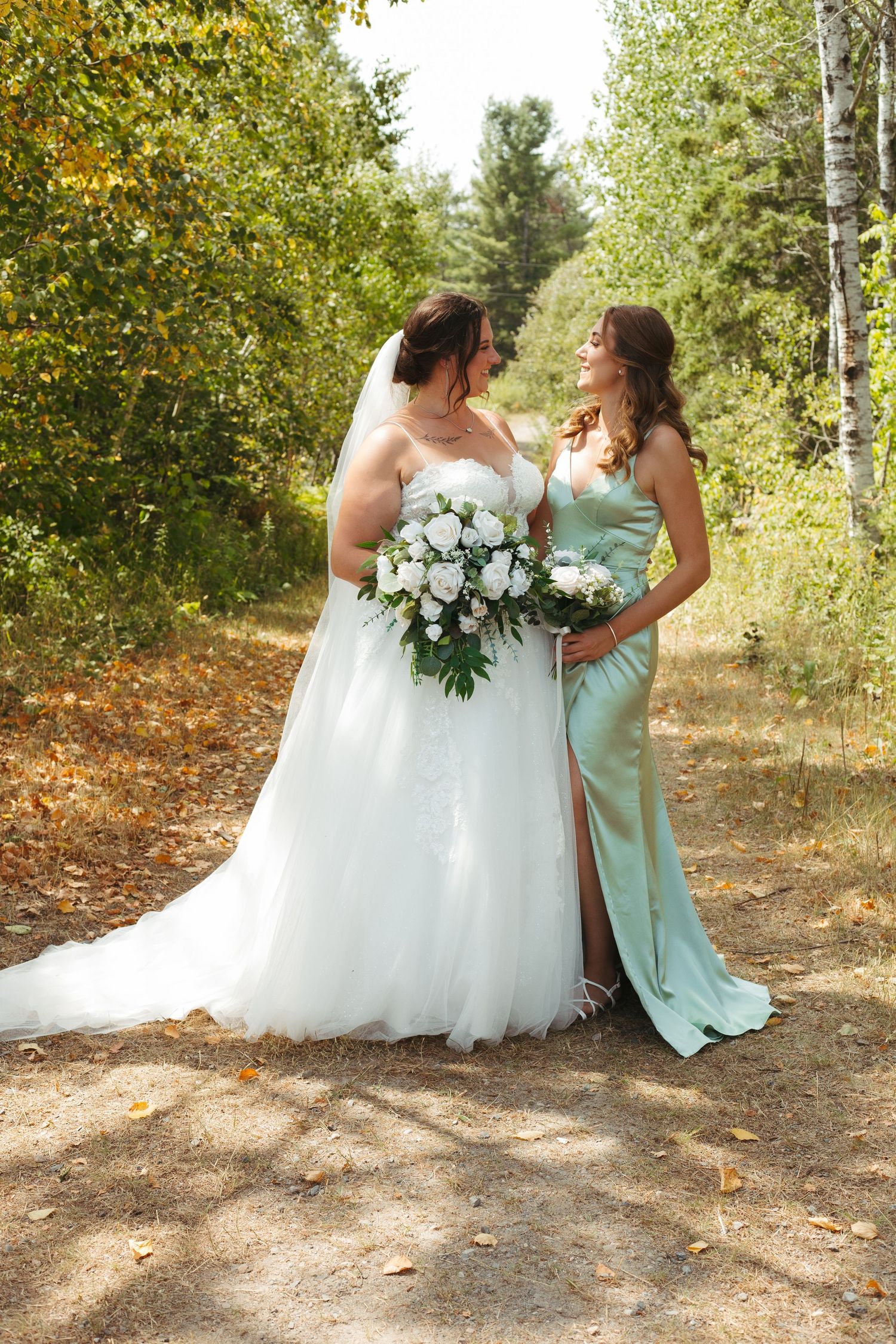 Bride in white ball gown and bridesmaid in mint green dress holding bouquets in a forest setting.