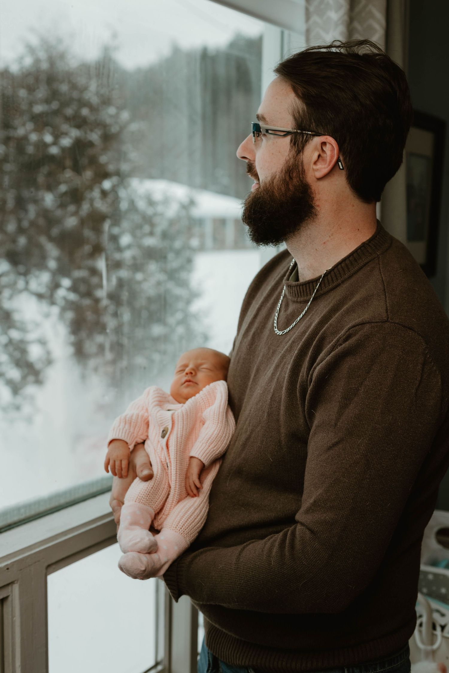 A bearded father in a brown sweater holds his newborn baby while gazing out a snowy window.