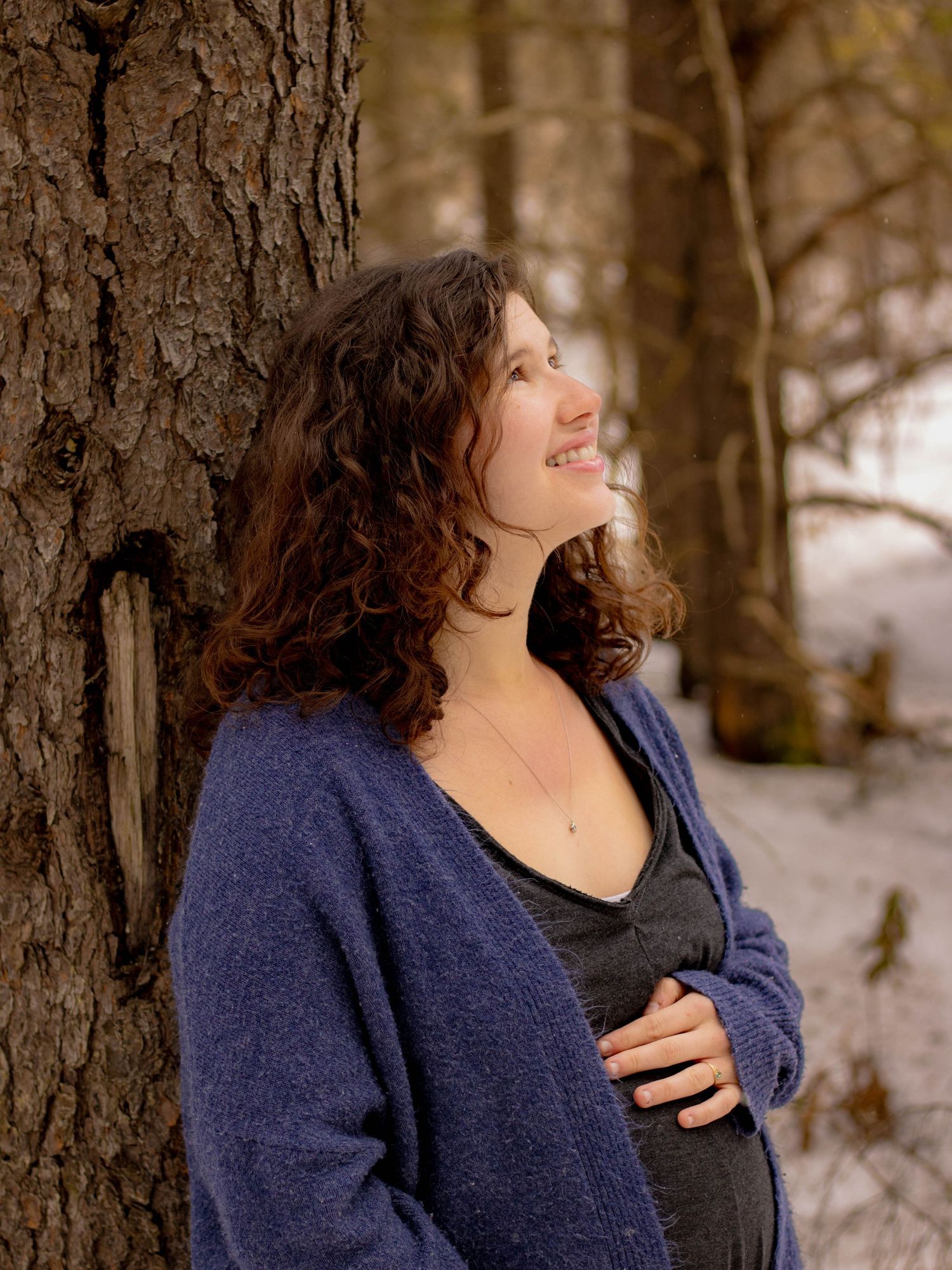 Smiling pregnant woman in blue cardigan leaning against tree in snowy winter forest.