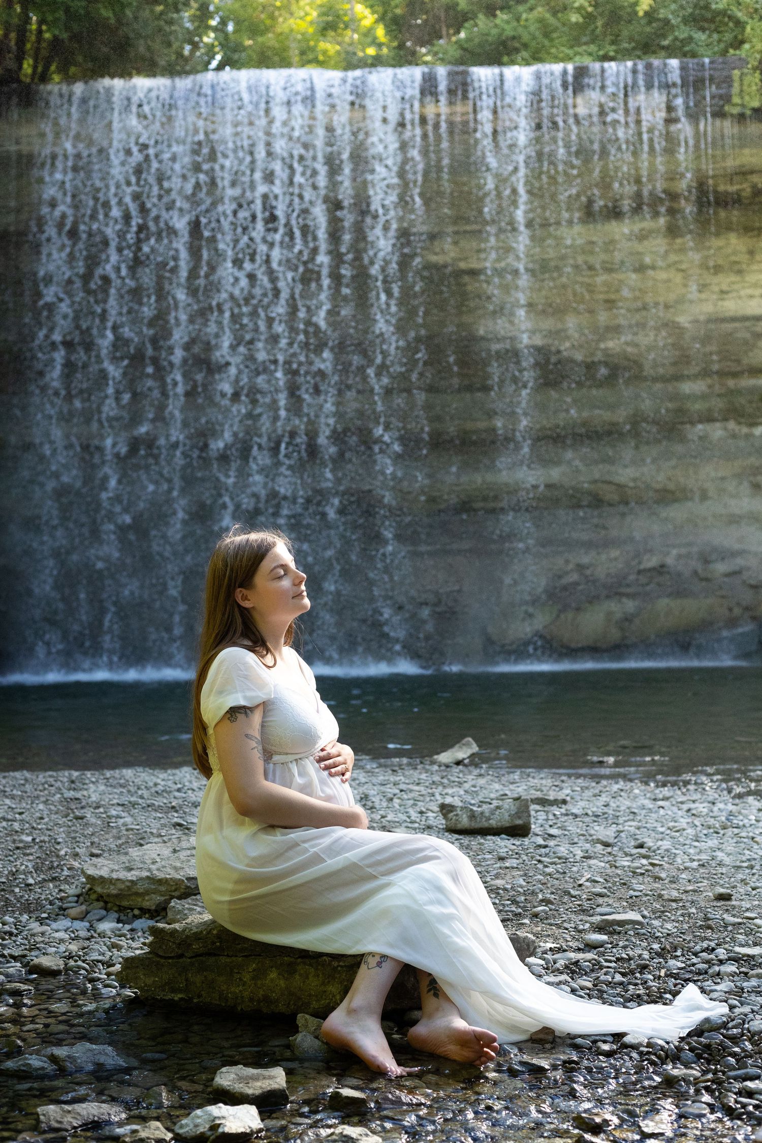 Pregnant woman in white dress sitting on rocks near a waterfall, enjoying the outdoors.