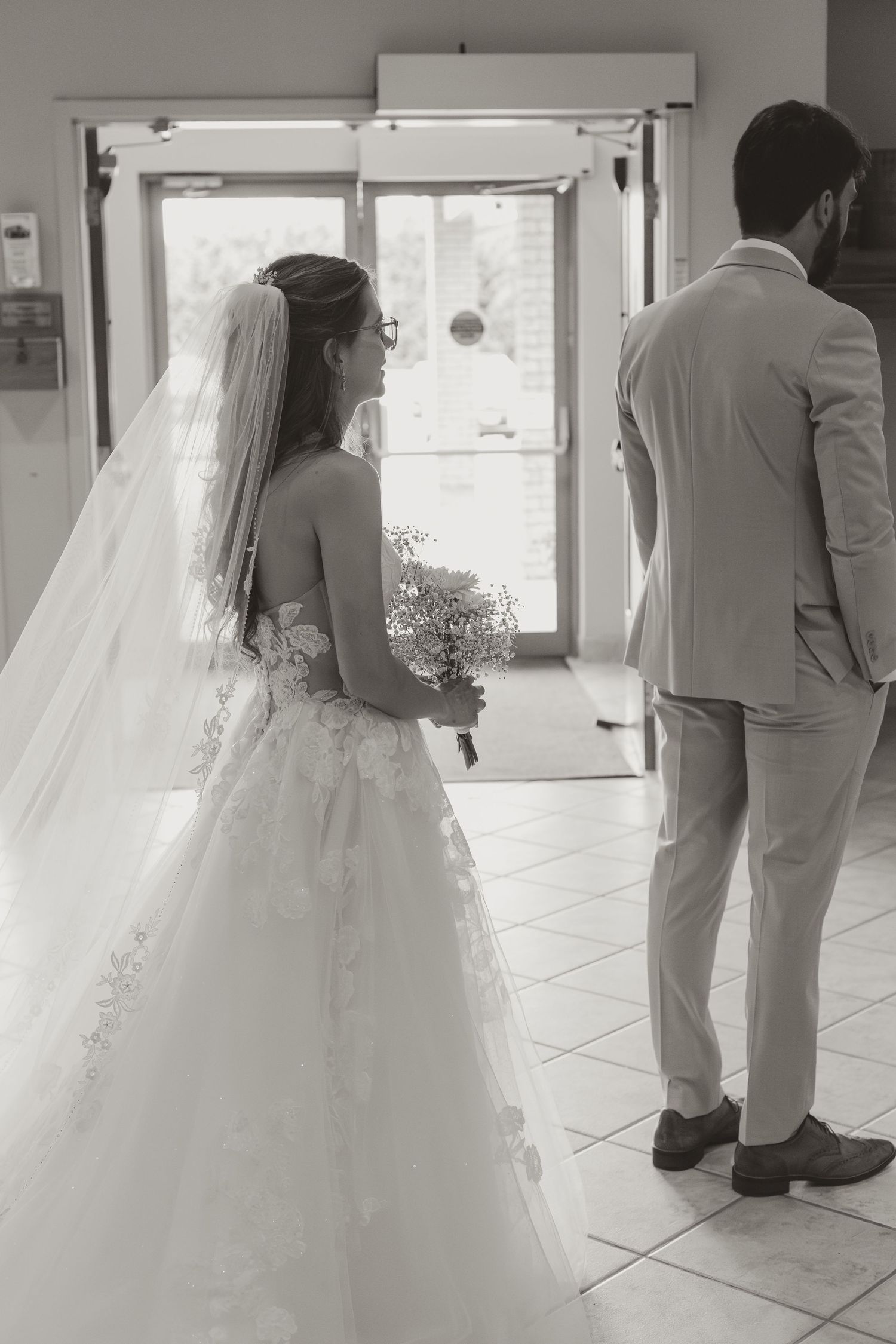 Black and white photo of bride holding bouquet and groom standing together near church entrance doors.