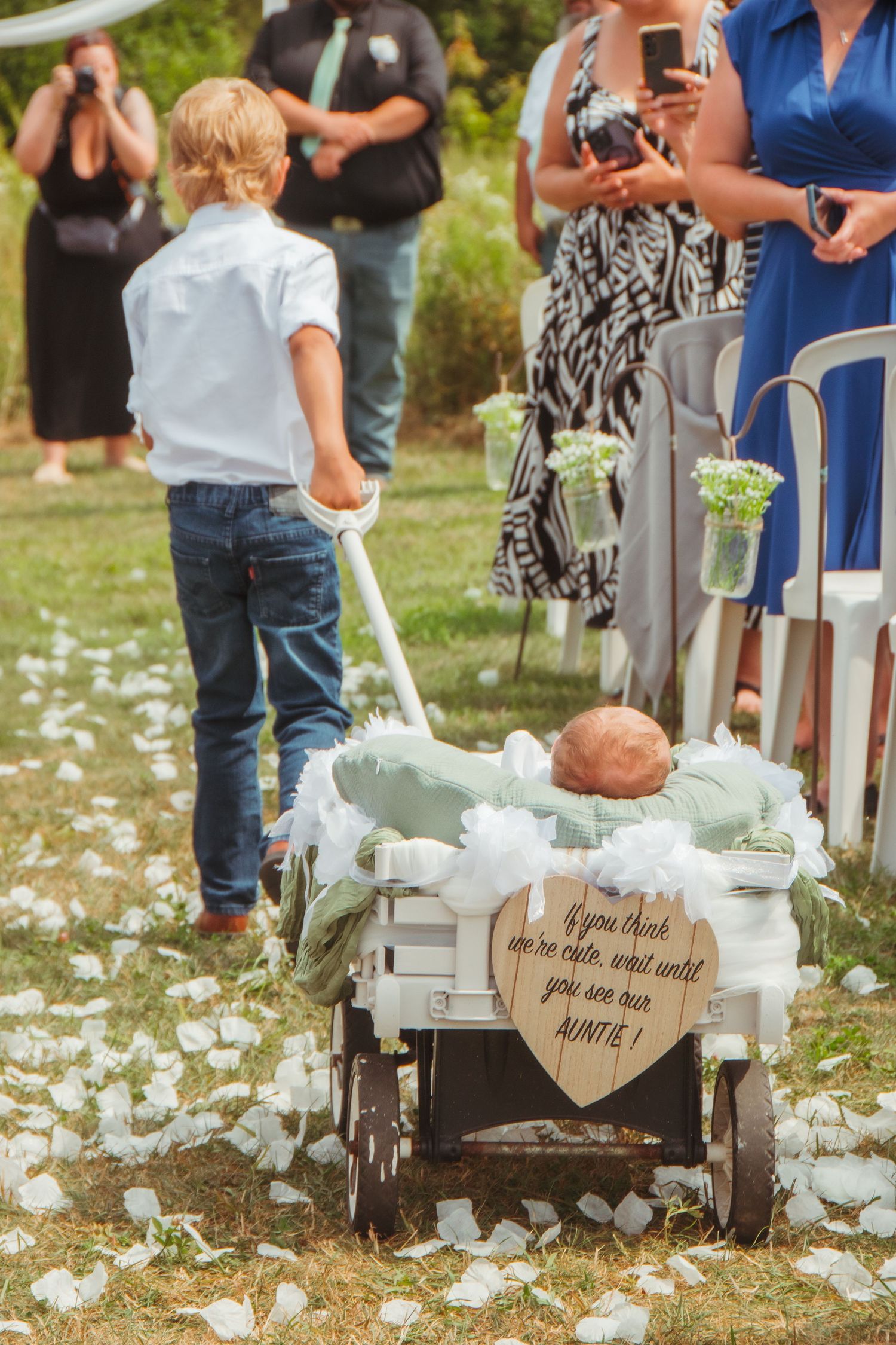 Boy pulls wagon with baby at wedding, sign reads 'wait until you see our Auntie', white petals line the aisle.