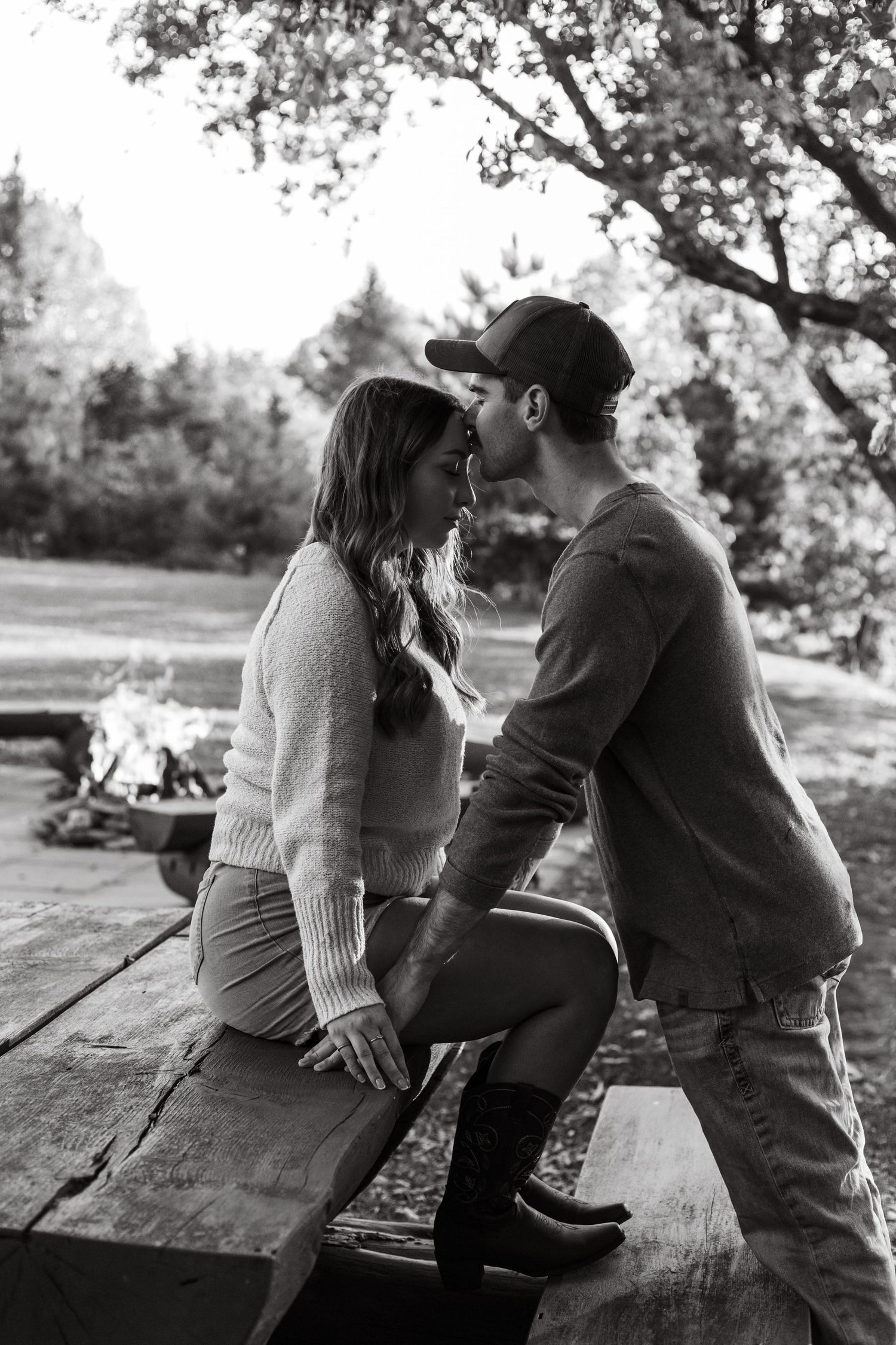 A couple leans in close together on a wooden bench with a fire glowing in the background.