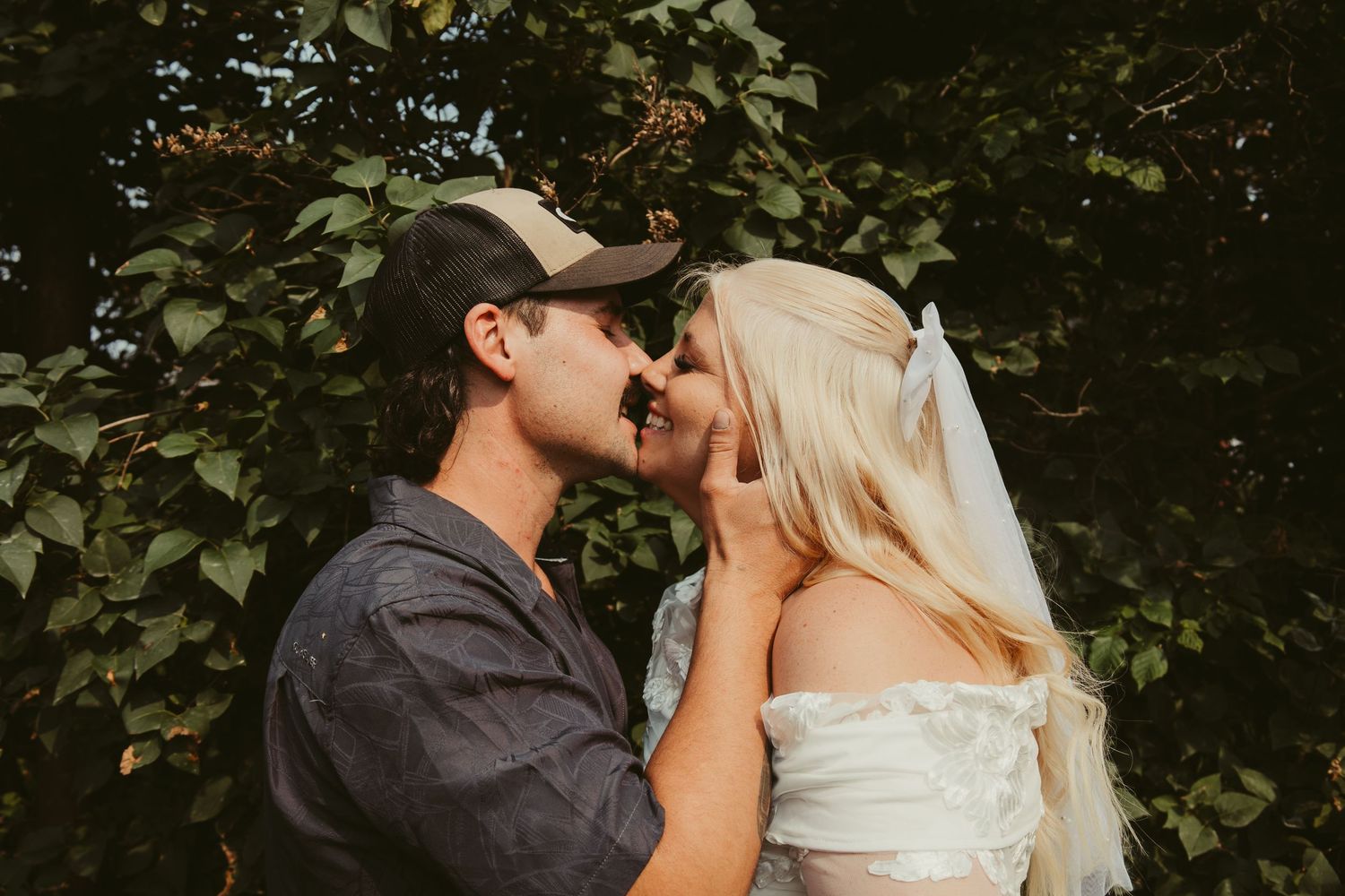 Bride and groom sharing a romantic kiss outdoors, bride wearing a veil and groom in a casual cap.