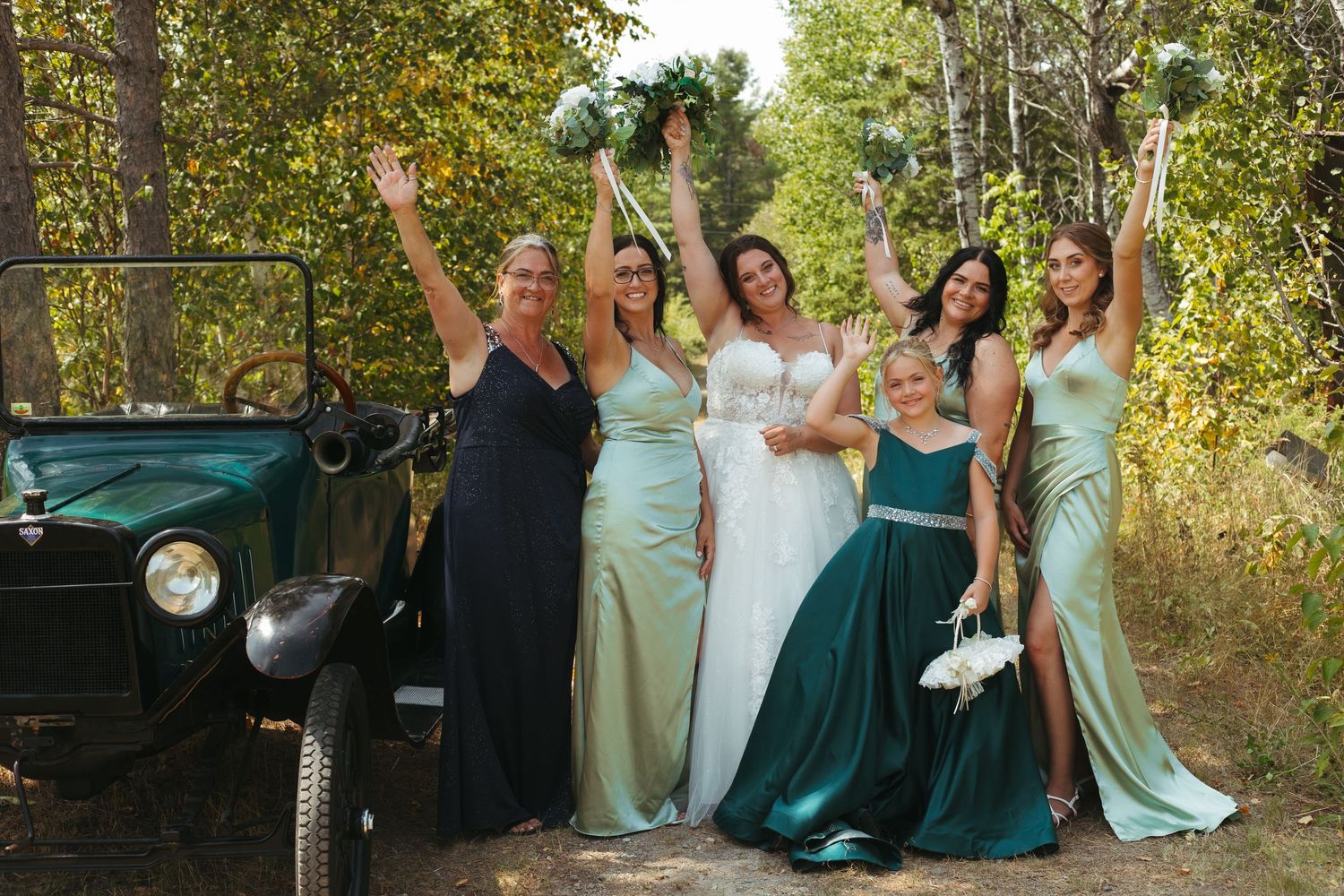 Bride and bridesmaids in green dresses celebrating beside vintage car outdoors.