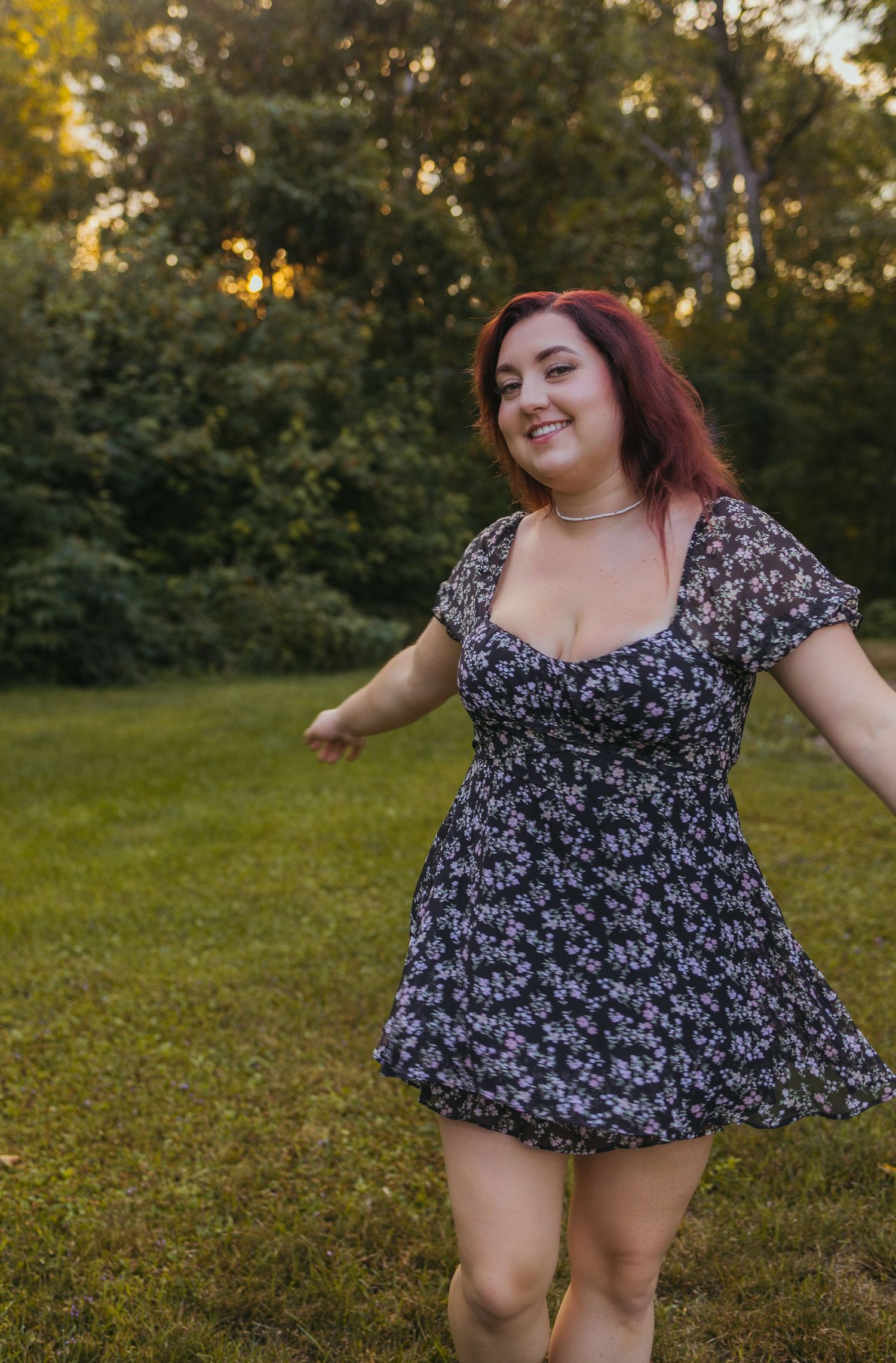 Woman in floral mini dress smiling and twirling outdoors on green grass with trees in background.